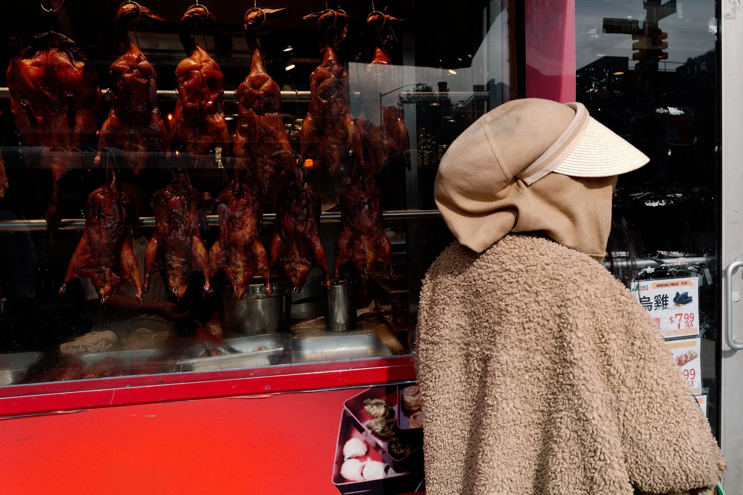 A hooded passerby pauses at a Chinatown storefront, facing rows of roasted birds behind glass. Texture, reflection, and display compress into a brief encounter between figure and window.