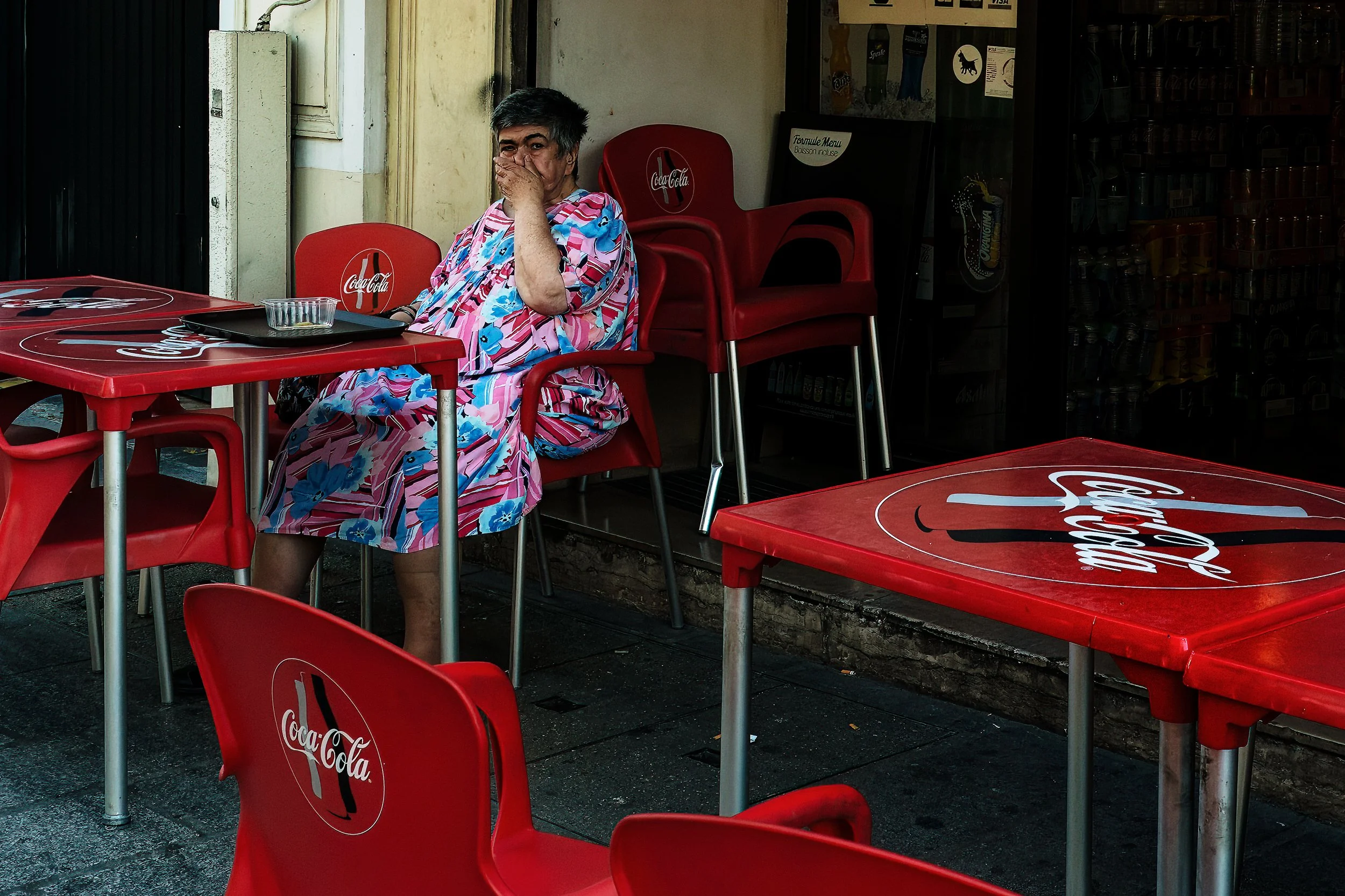 A solitary figure sits among empty café tables, framed by red chairs and the repeated marks of everyday commerce. Her quiet gesture gives the scene a sense of pause, turning the space between tables into a small moment of reflection