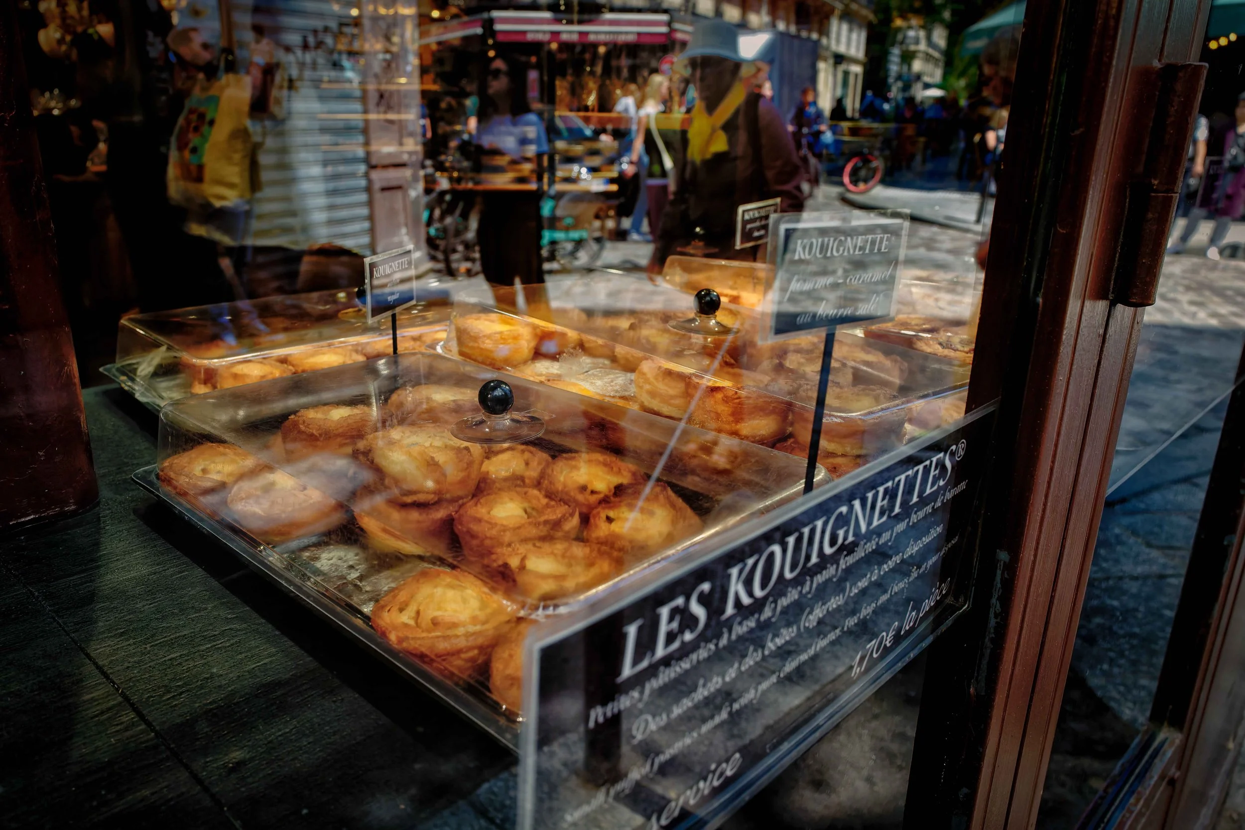 Street Pastry: Golden kouignettes sit beneath clear covers in a bakery window as pedestrians, bicycles, and the surrounding Paris street overlap in the reflected glass.