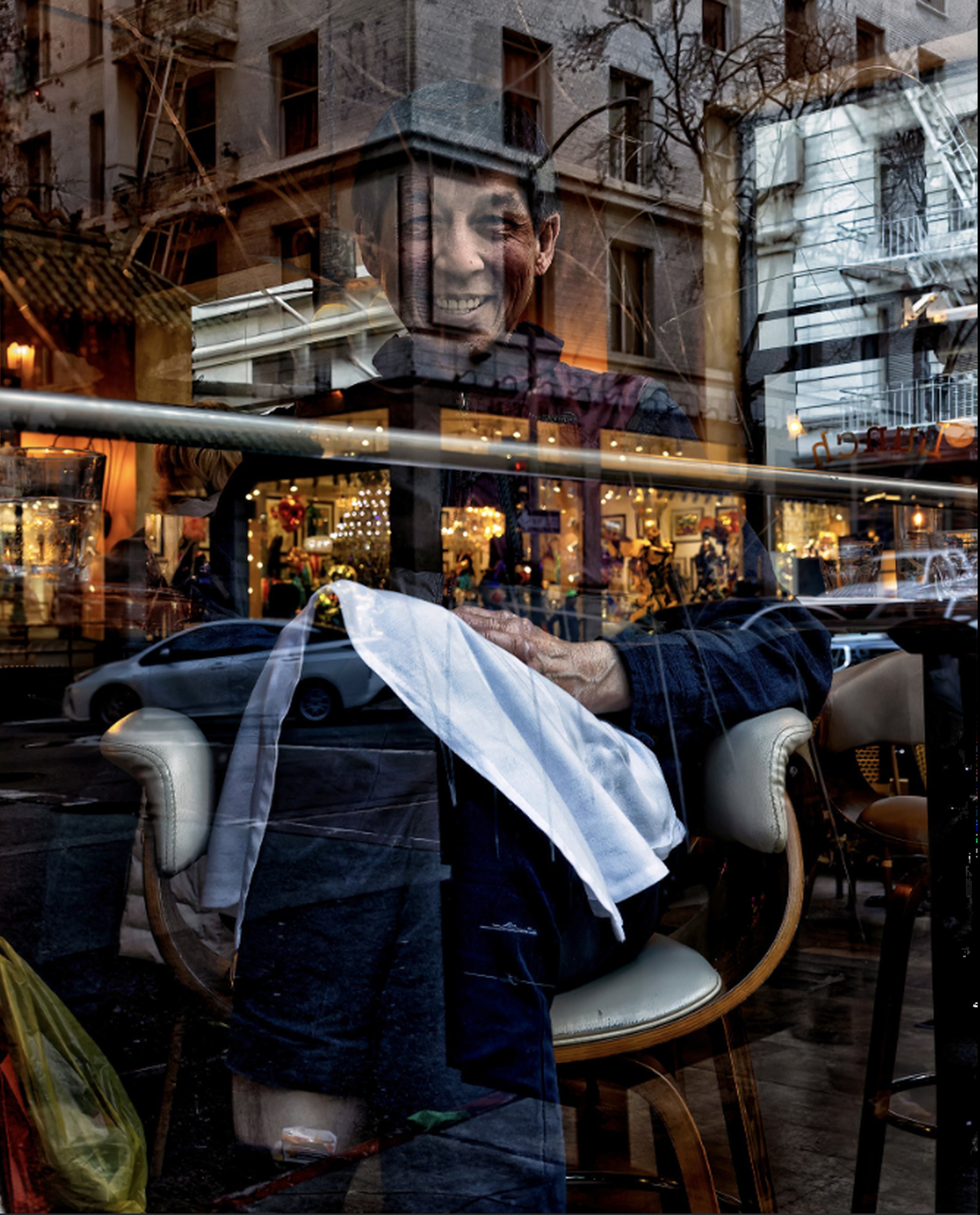 Smiling person holding a white cloth seen through a restaurant window, layered with reflections of buildings, cars, lights, and city street activity.