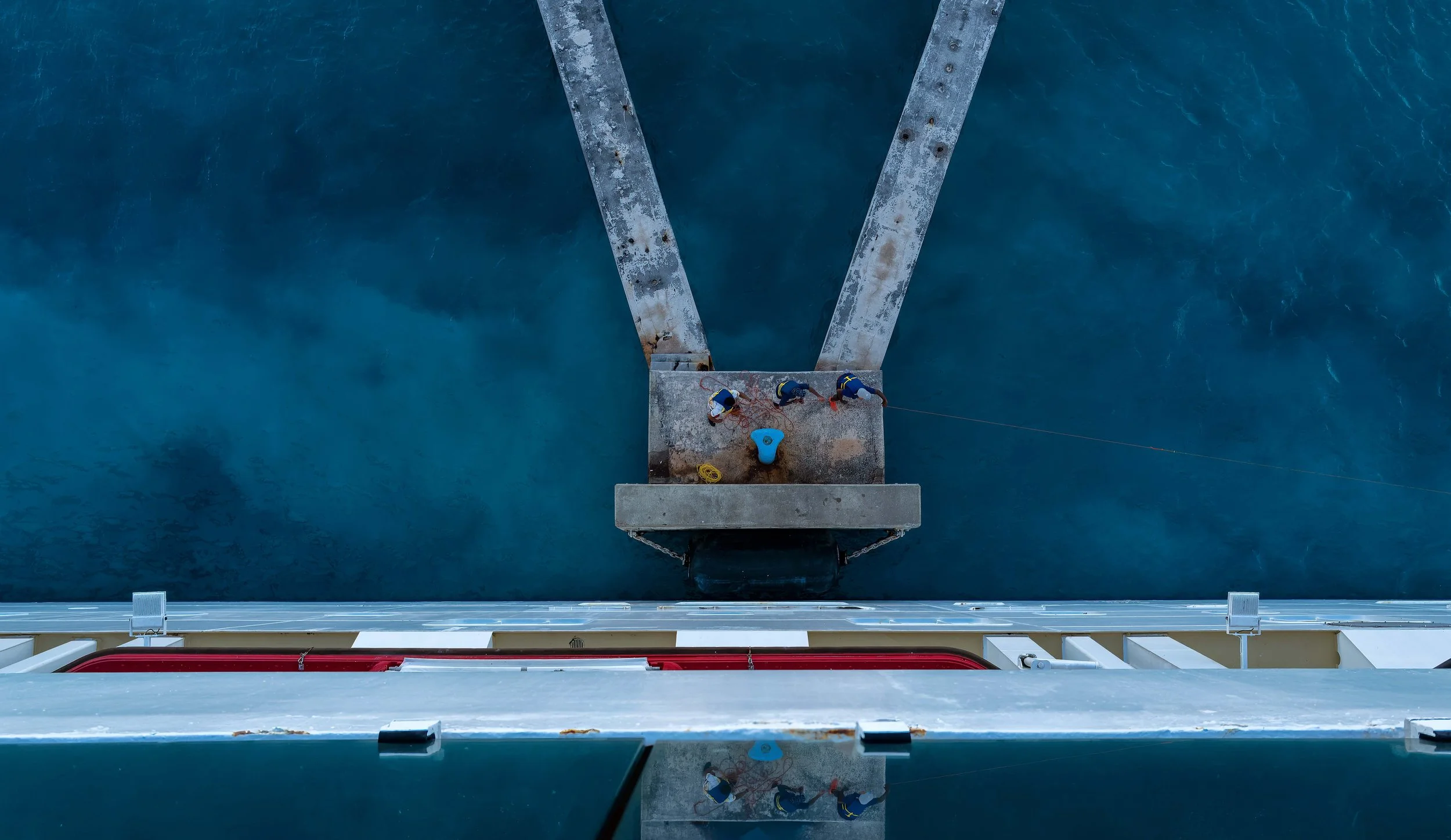 Workers seen from above on a small platform over blue water, with bridge supports and reflections creating a symmetrical composition.