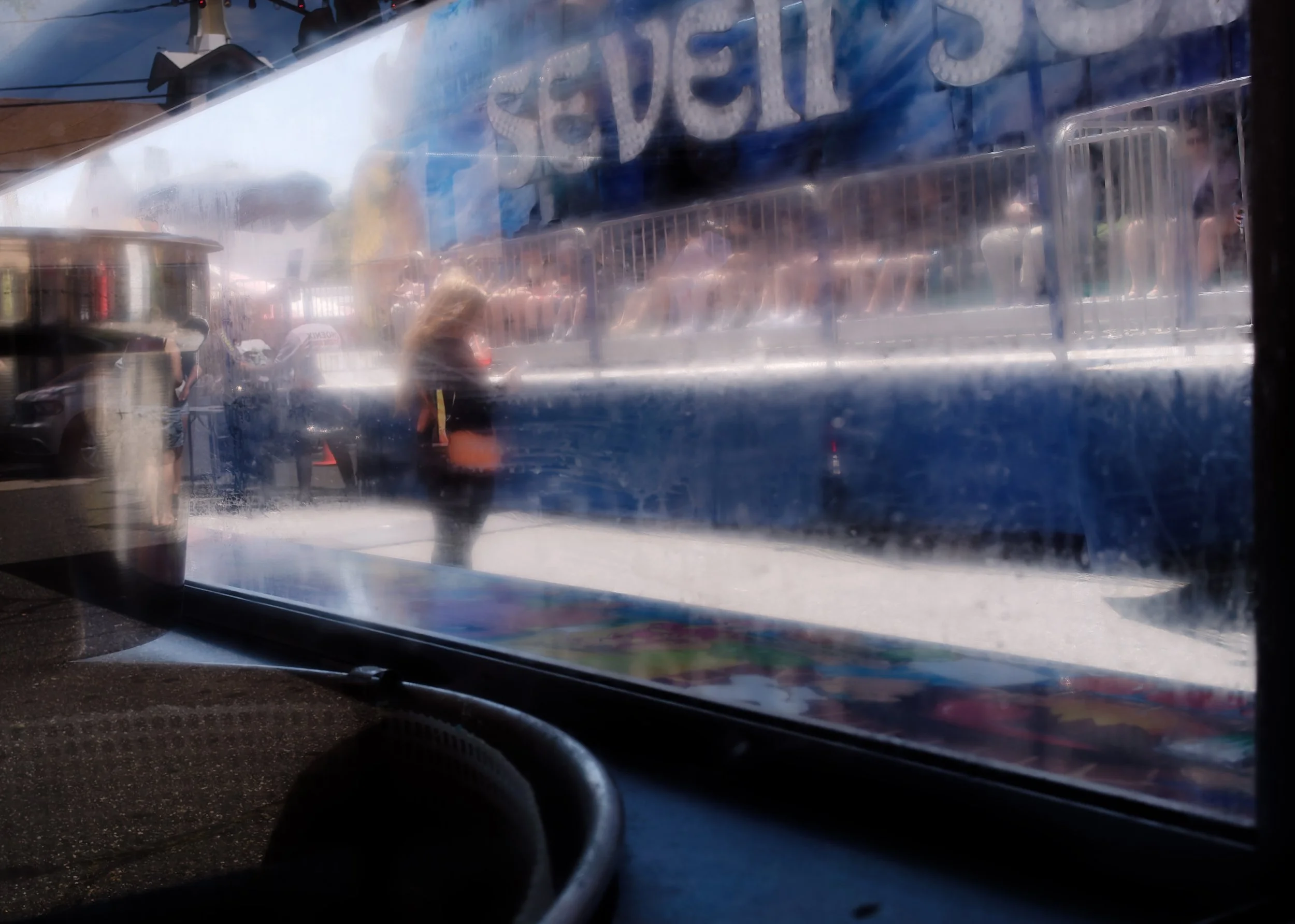 Blurred figure passing outside a window, seen through layered glass reflections, blue signage, street barriers, and a dim interior foreground.