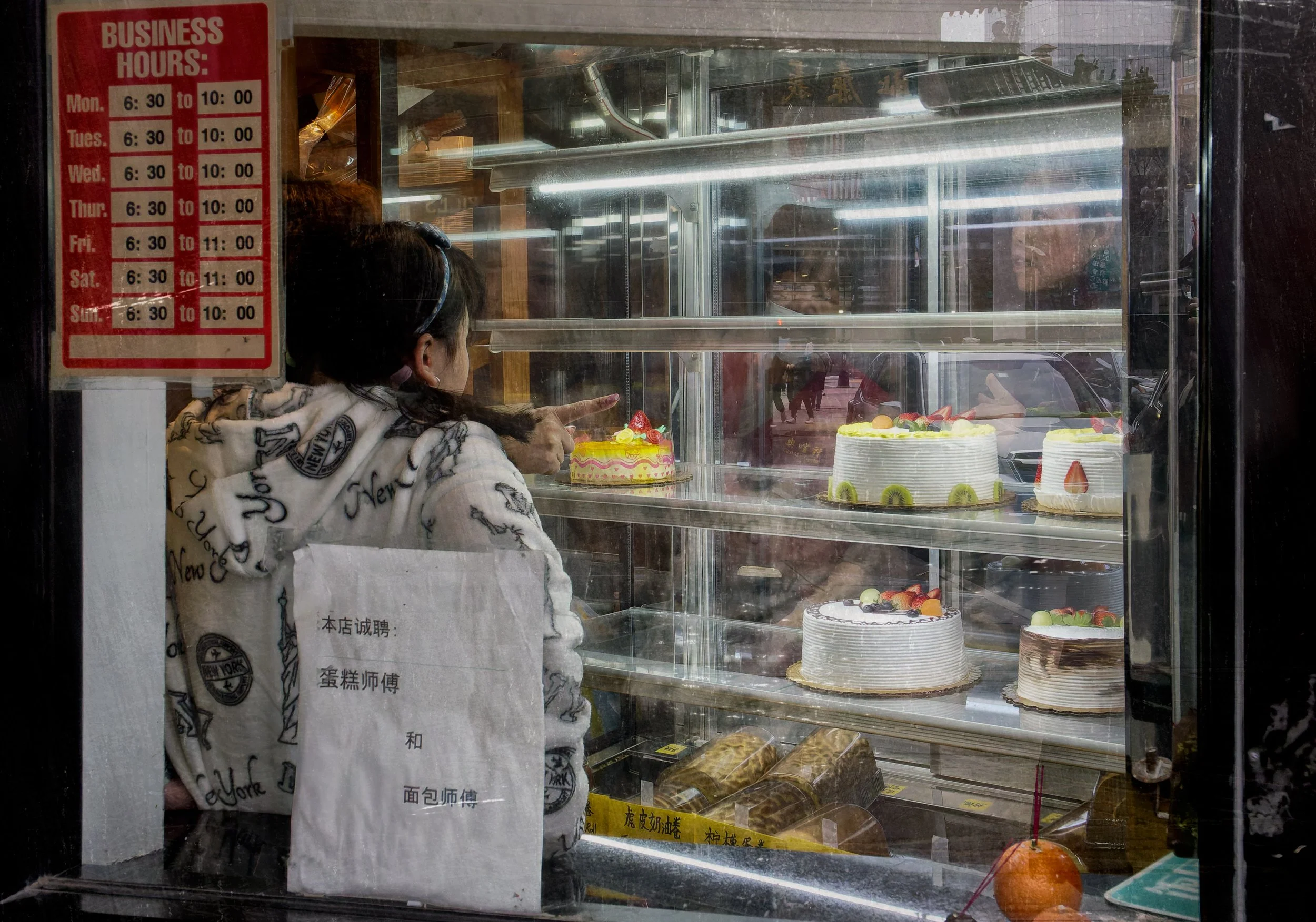 Person looking into a Chinatown bakery display case filled with cakes, layered with window reflections, business hours signage, and street details.