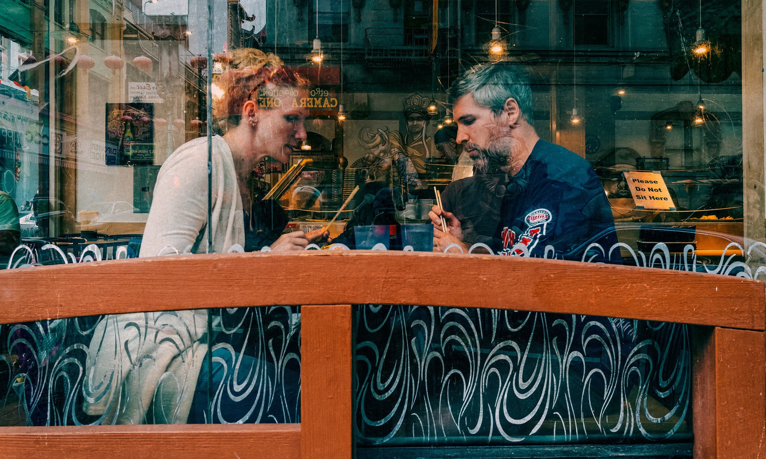 Two diners eating noodles inside a Chinatown restaurant, seen through a window layered with street reflections, signage, hanging lights, and patterned glass.