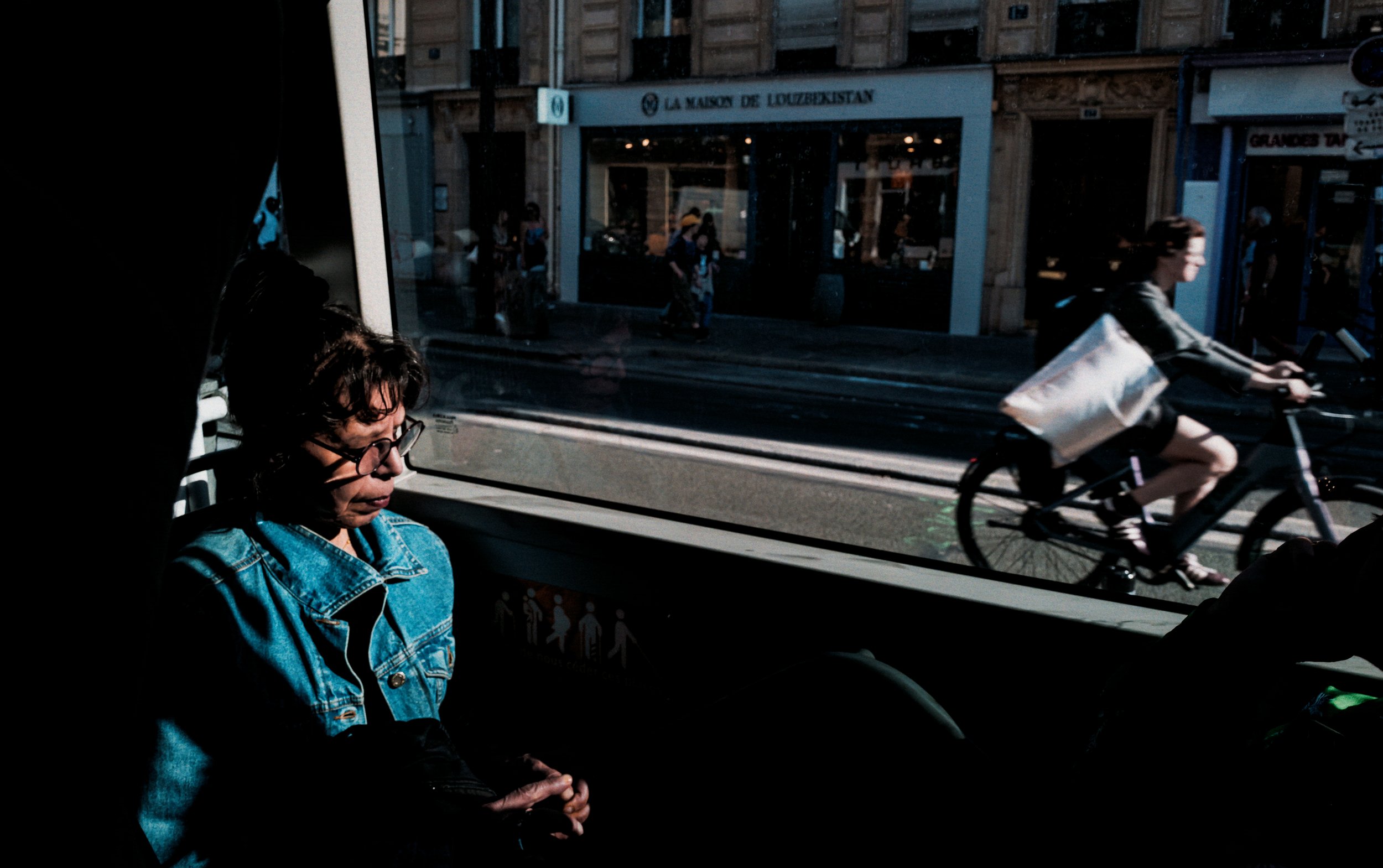 Woman seated in deep shadow inside a bus or tram, with a cyclist and city street visible through the window beside her.