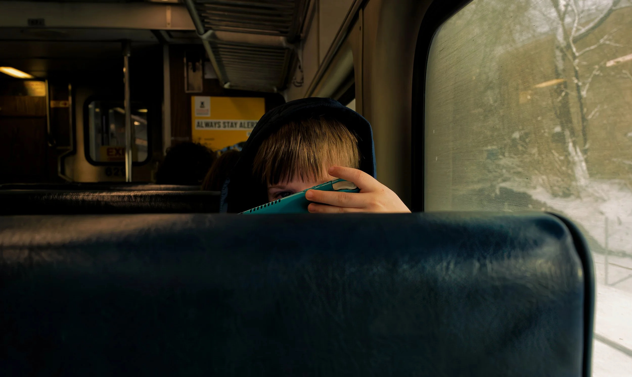 Child peering over a train seat while holding a tablet, framed by a dim train interior and window light.