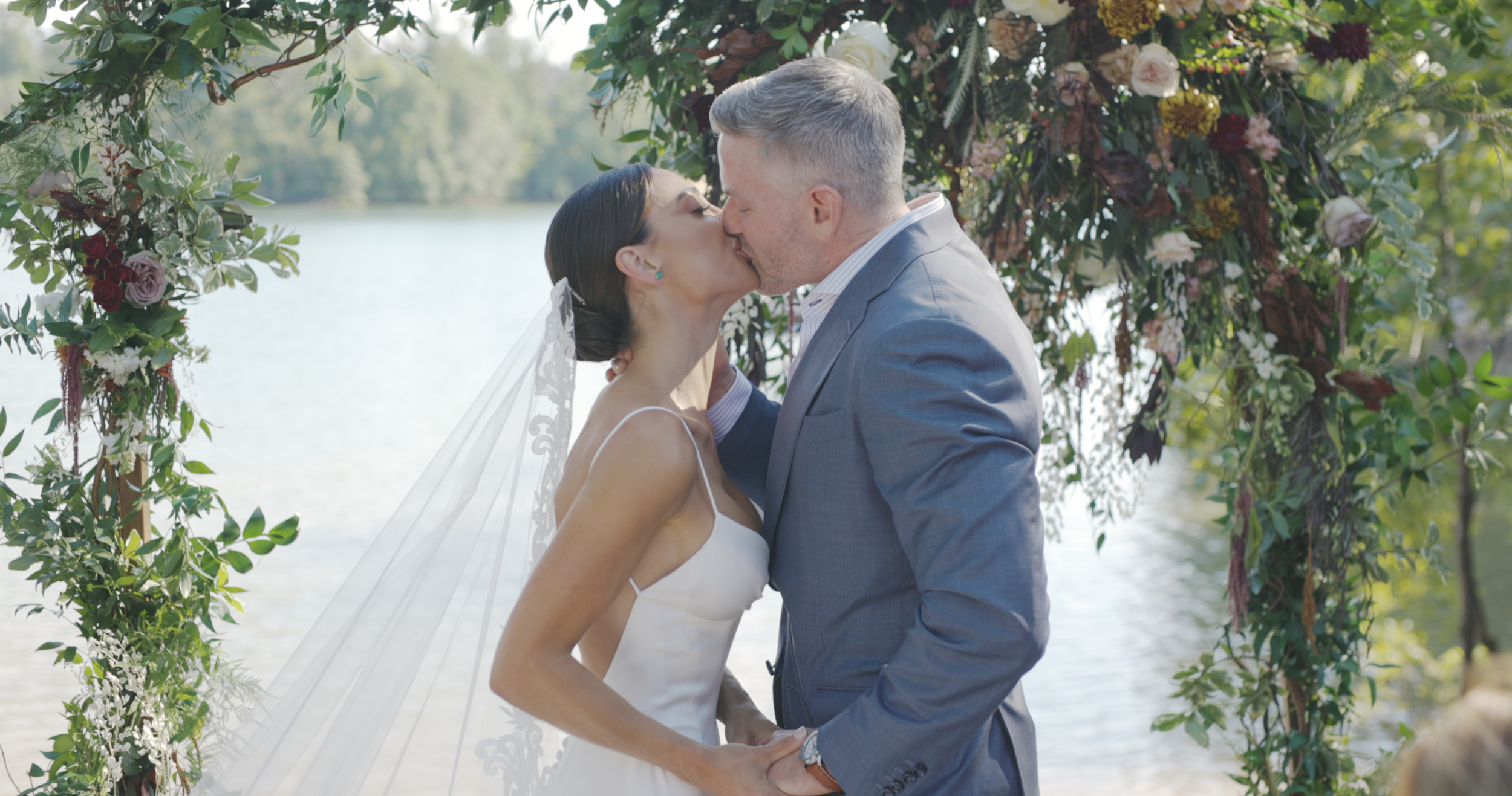 Bride and groom kissing under a floral wedding arch beside a lake, wedding film still, Greenville SC