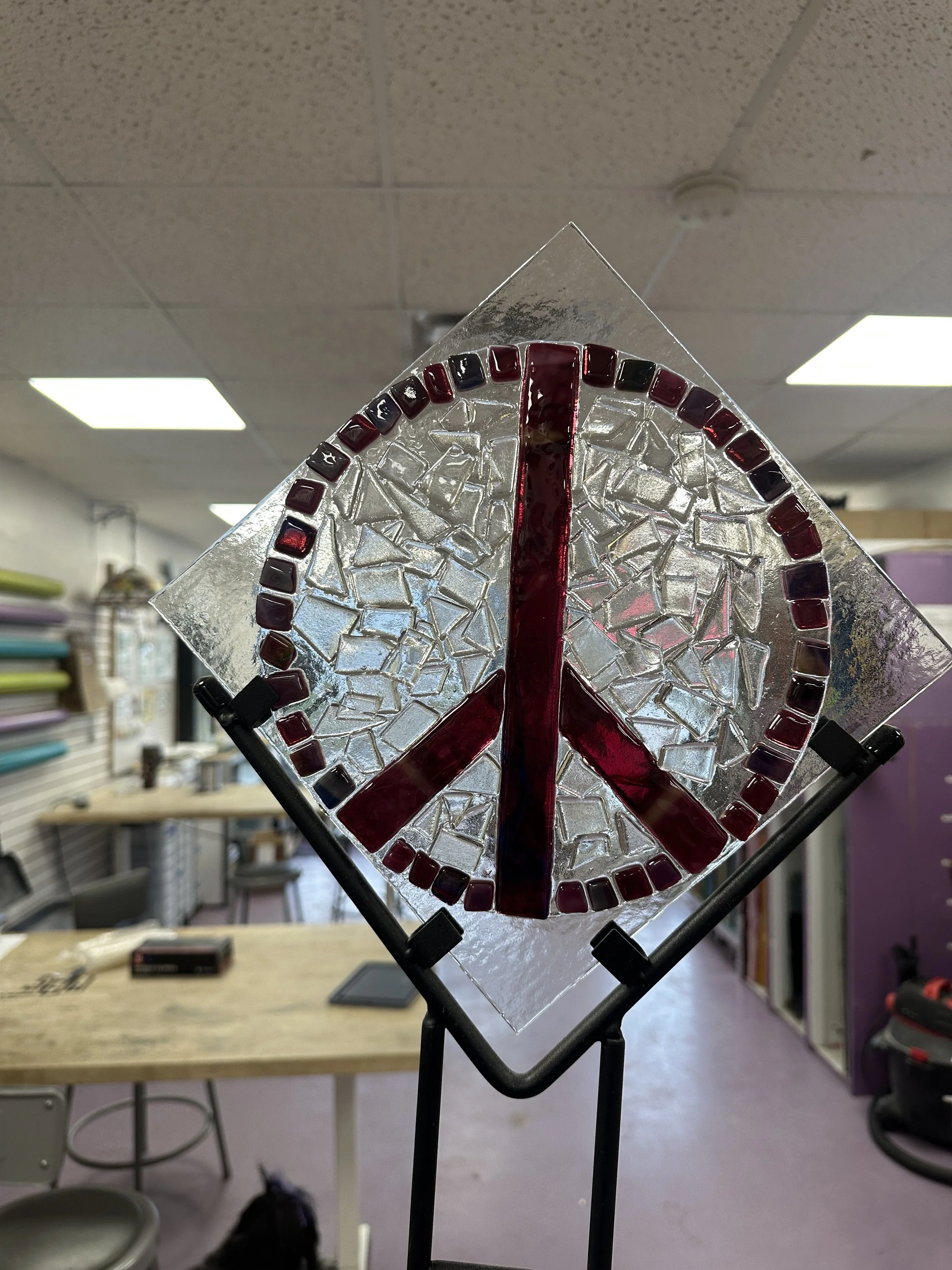 two students standing smiling by their work table where they are assembling fused glass garden stakes using sheet glass, stringers of glass, and other fusing materials. above them is a stained glass lamp that stern glass works repaired. 