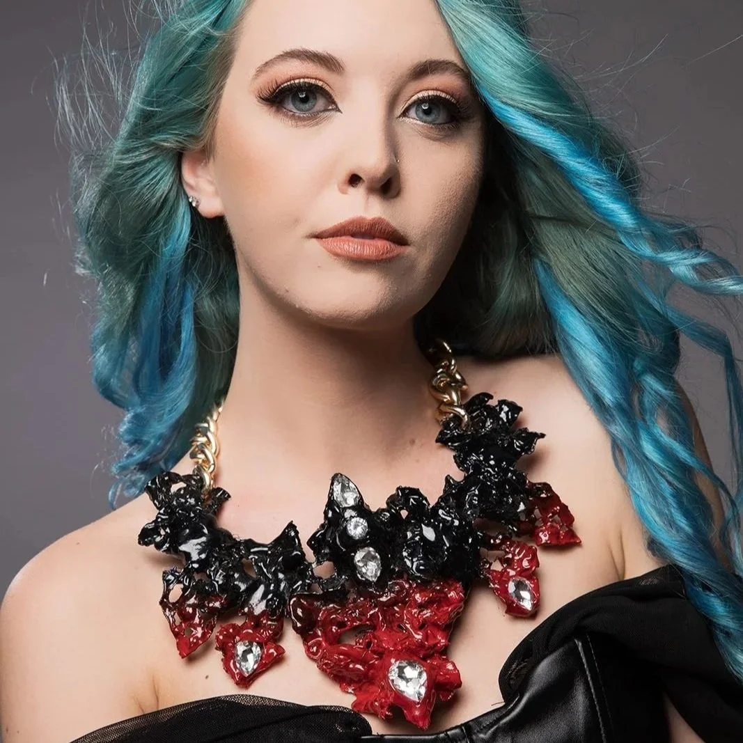 Portrait of a woman with blue and green wavy hair, wearing makeup, a large black, red, and clear gemstone necklace, and a black top.