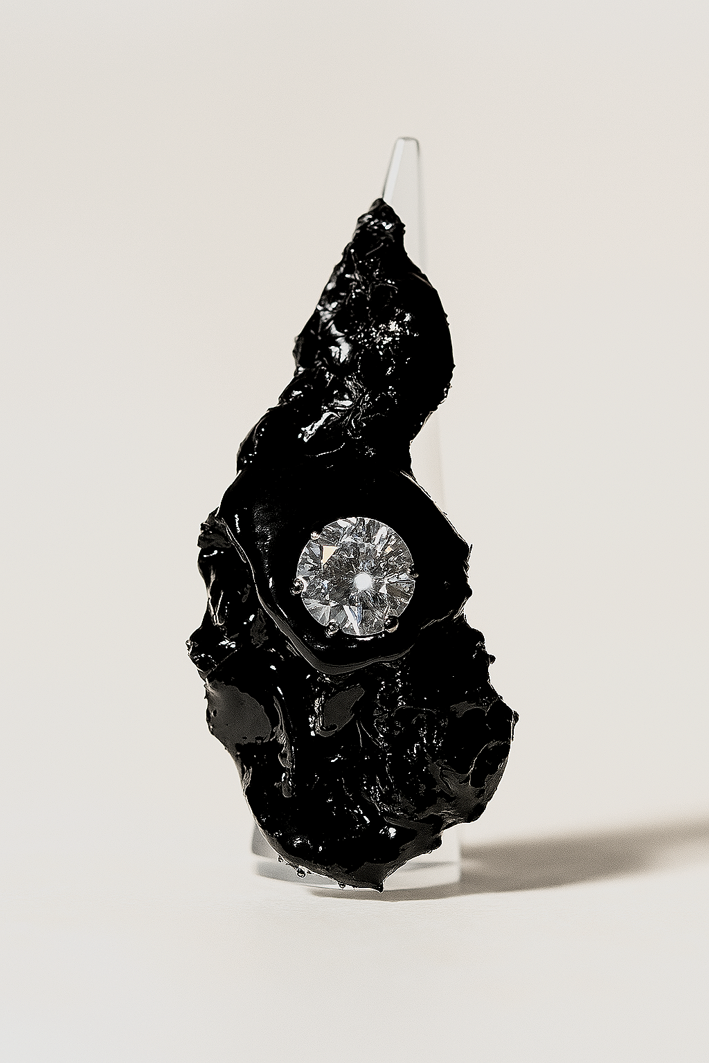 An abstract sculpture made of shiny black material with a large, clear, faceted gemstone embedded in it, displayed on a white background.