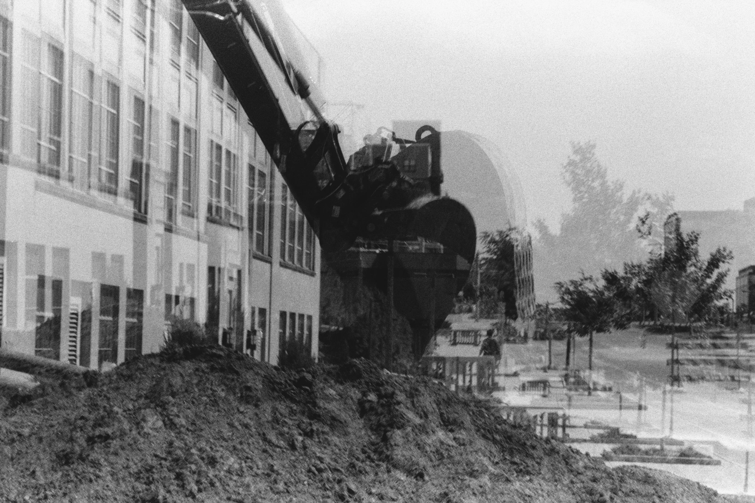 A black-and-white double exposure shows an excavator over a dirt pile outside UIowa’s Engineering Building, with trees and structures faintly visible.