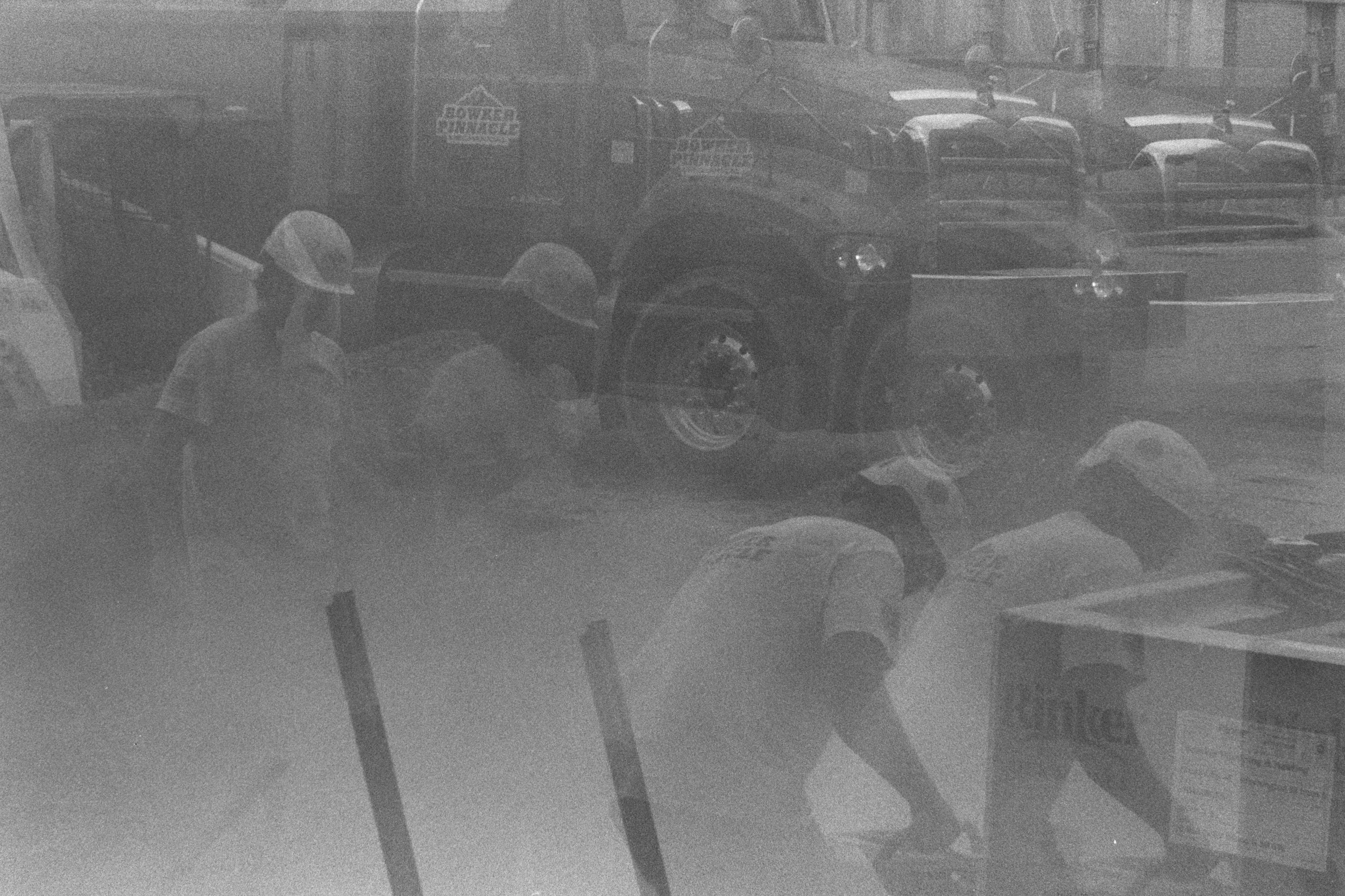 A double-exposed image shows two construction workers in hard hats, one operating machinery and the other walking toward him, with a truck behind them.