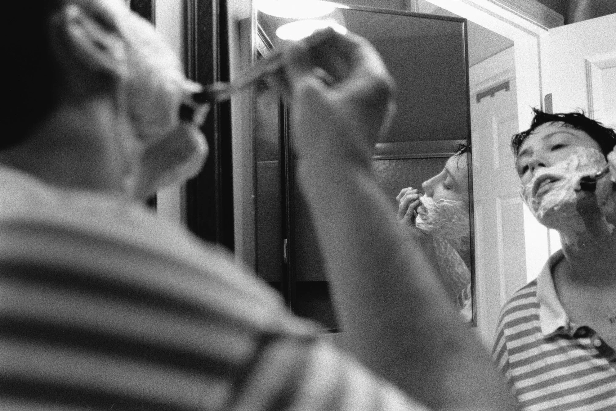 A self-portrait shows a person shaving, out of focus in the foreground, with two in-focus mirror reflections in a striped polo.
