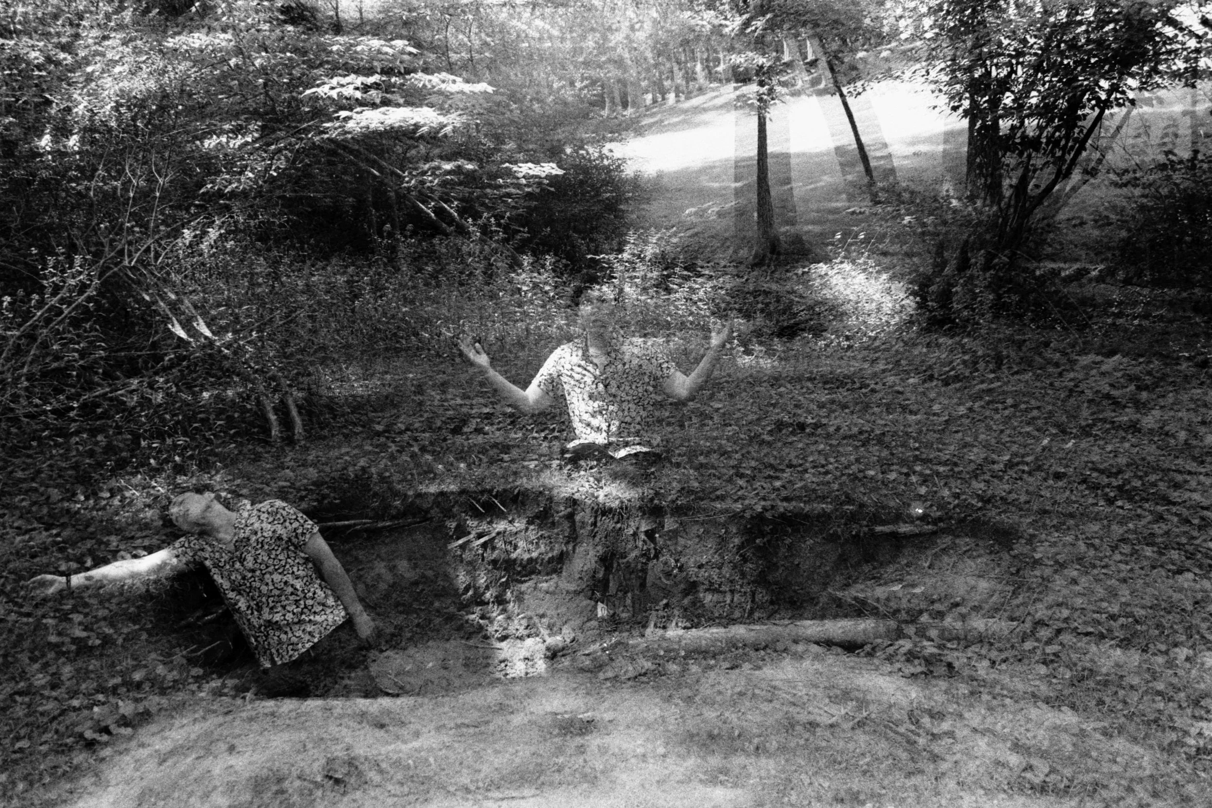 A black-and-white double-exposure self-portrait shows one figure lying in a hole in the woods and another seated at its edge with raised palms.
