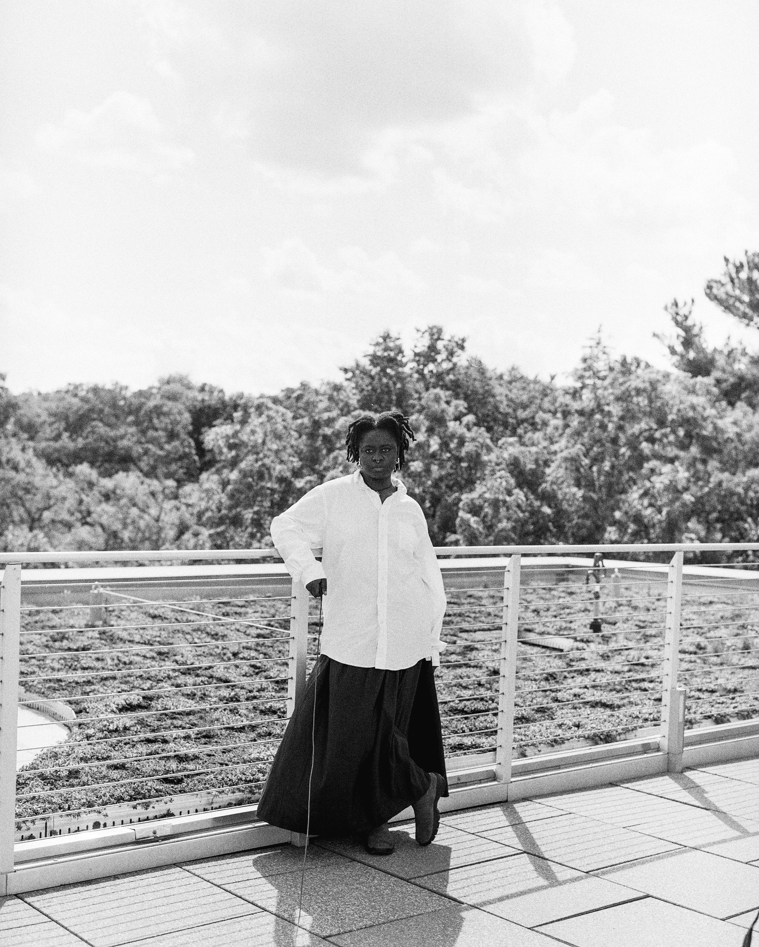 Esé stands on the Visual Arts Building roof in a white button-down, long black skirt, and boots, holding a cable release and looking at the camera.