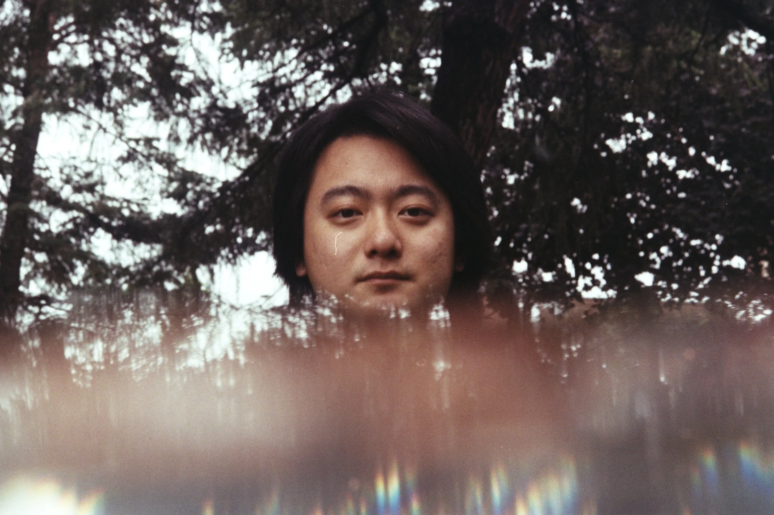 A dark-haired man from Handan, China, looks at the camera outdoors near Dubuque Street Hill, with foliage, a foreground prism, and a tear-like effect beneath one eye.