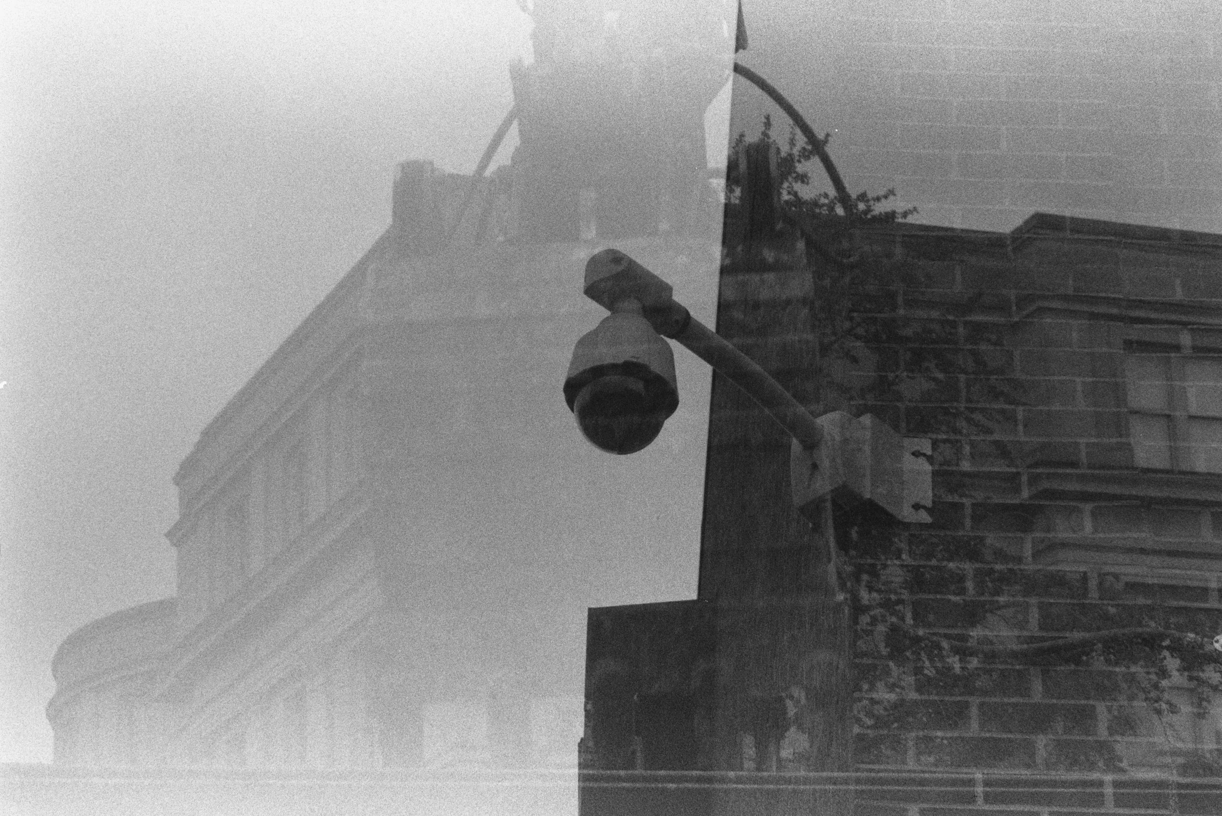 A surveillance camera is mounted on the corner of a brick building, with an institutional façade superimposed in the background.