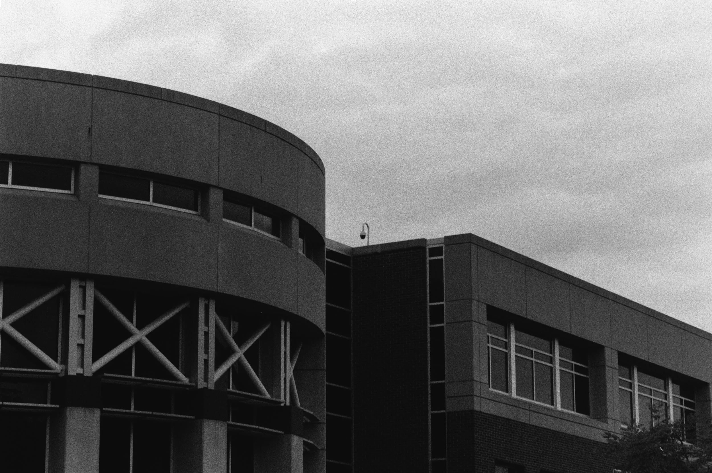 A surveillance camera sits between the curved and rectangular sections of a modern concrete building beneath a cloudy sky.