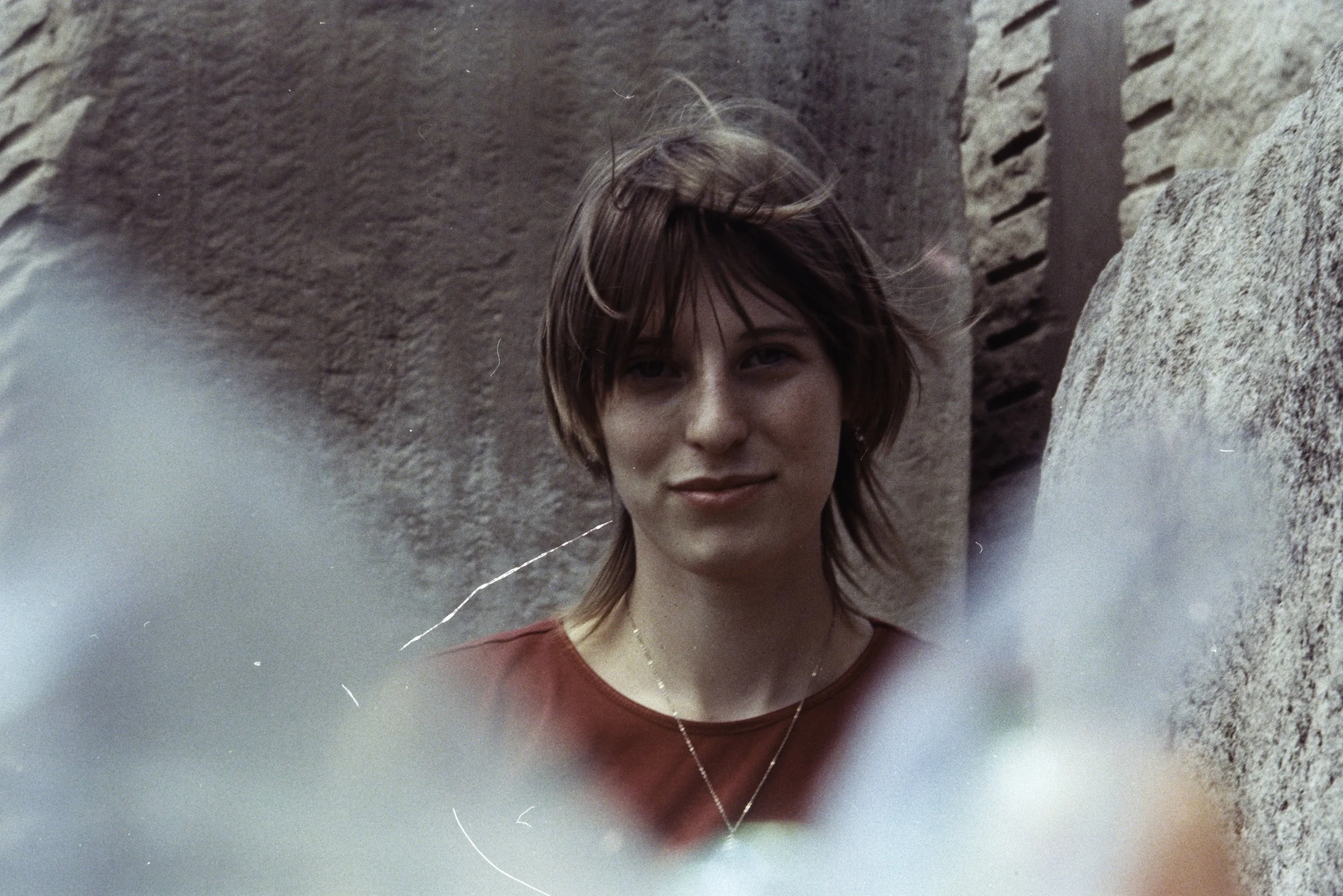 A young person with short brown hair, a red shirt, and a gold necklace looks at the camera in front of a stone structure, with a blurred foreground.