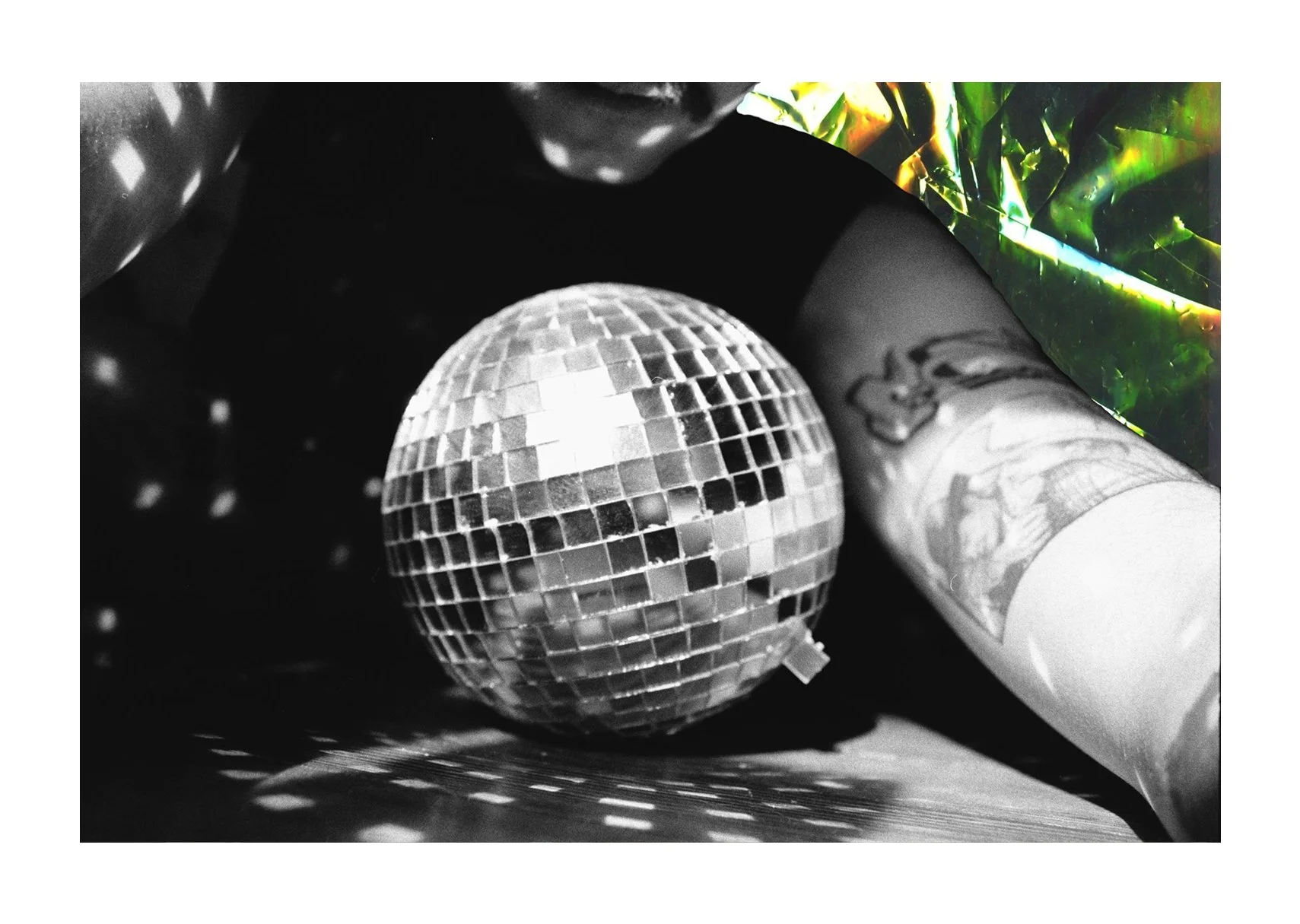 A black-and-white photo shows a disco ball on a wood floor, a tattooed figure leaning toward it, and cellophane in the top right.