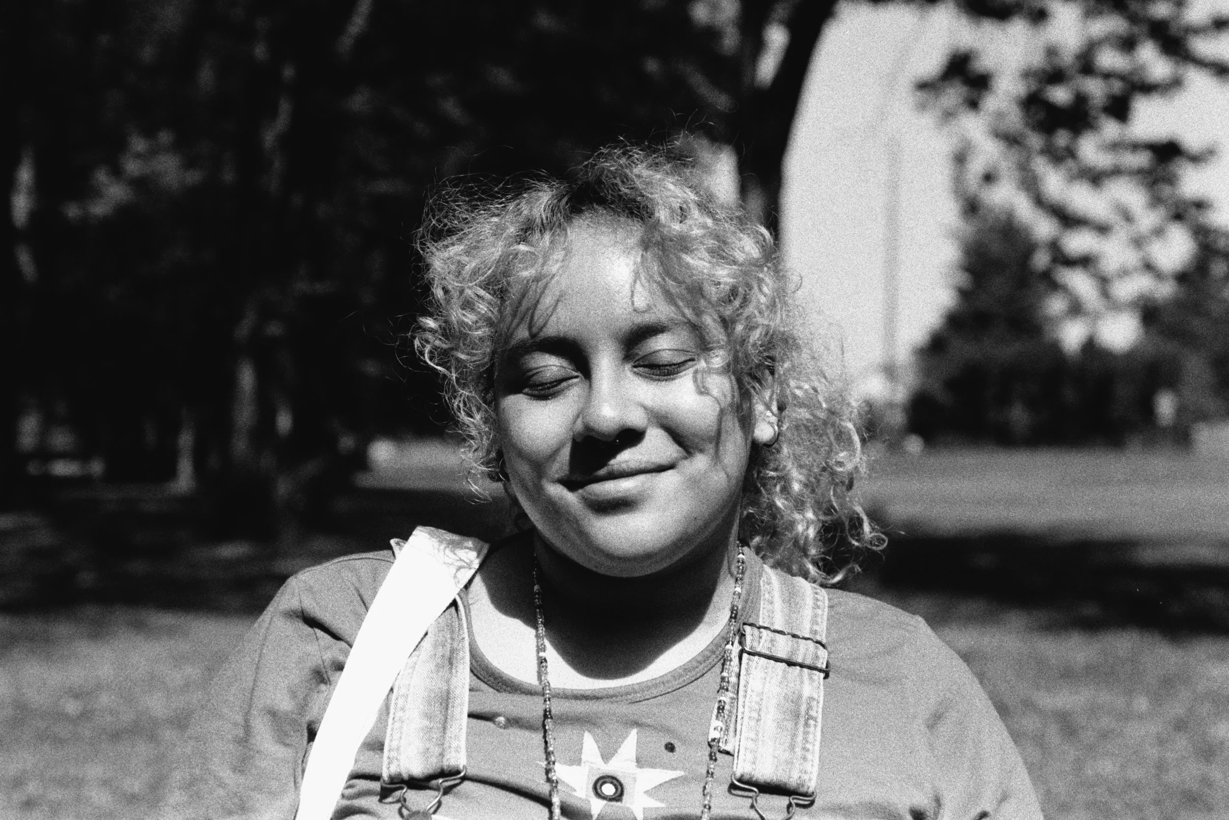 A black-and-white outdoor portrait of a smiling nonbinary person with curly hair and closed eyes.