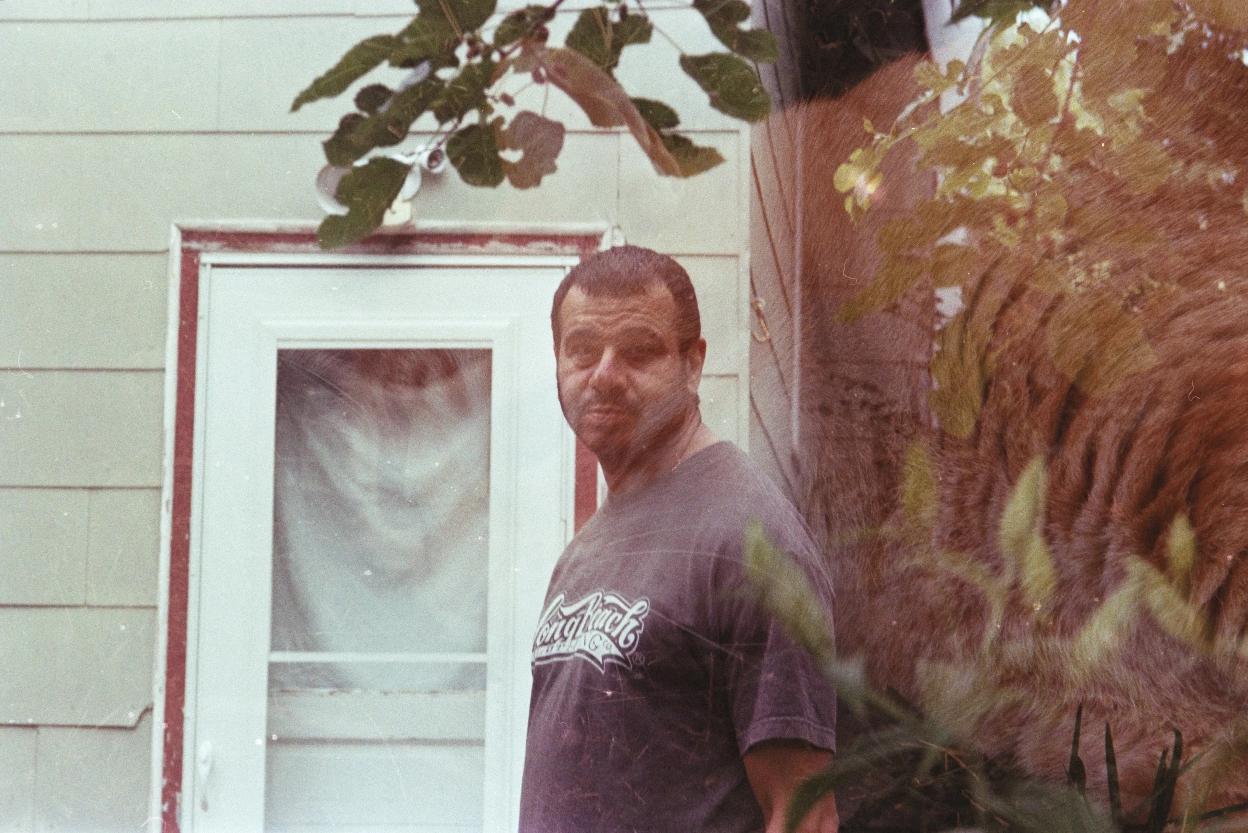 A young Egyptian man in a dark T-shirt stands by a white door with red trim, with a double exposure of his cat Cuddles resting in grass.