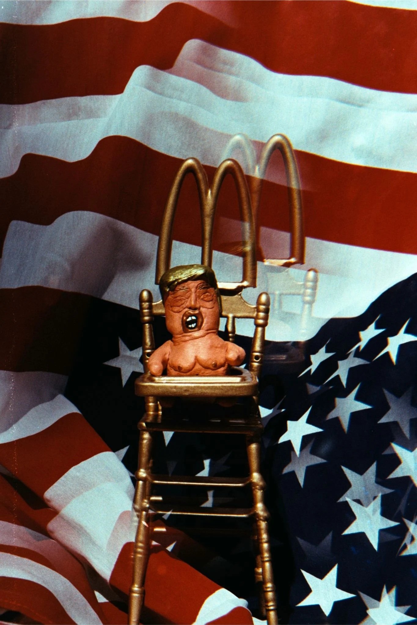 A color photo of a clay sculpture of DJT as a raging baby in a gold toy throne with McDonald’s arches, against an upside-down U.S. flag, doubled by a prism.