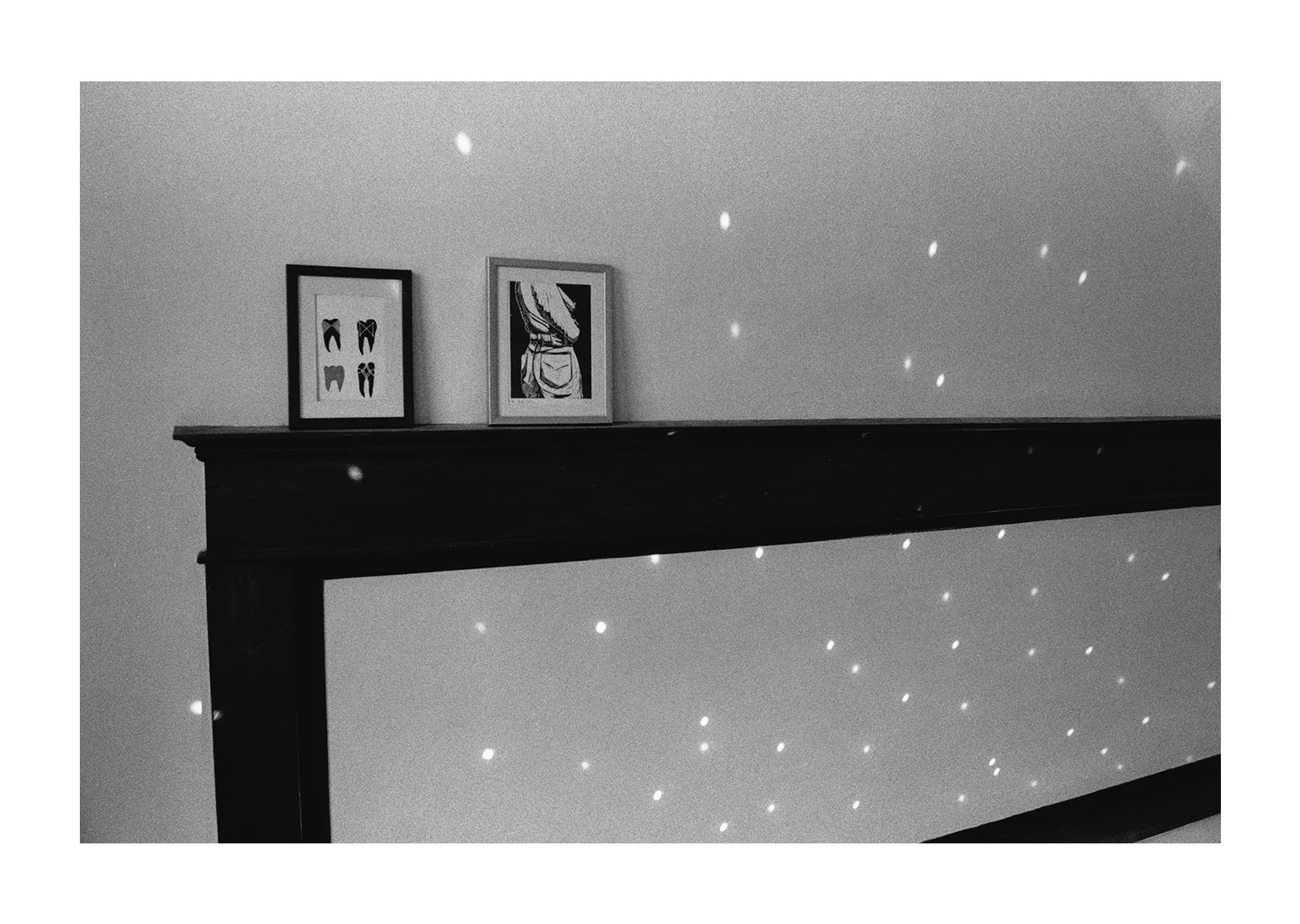 n black & white, many dots of light reflected off of a disco ball (out of frame) cover a wall where two framed art prints rest on a built-in wooden ledge.