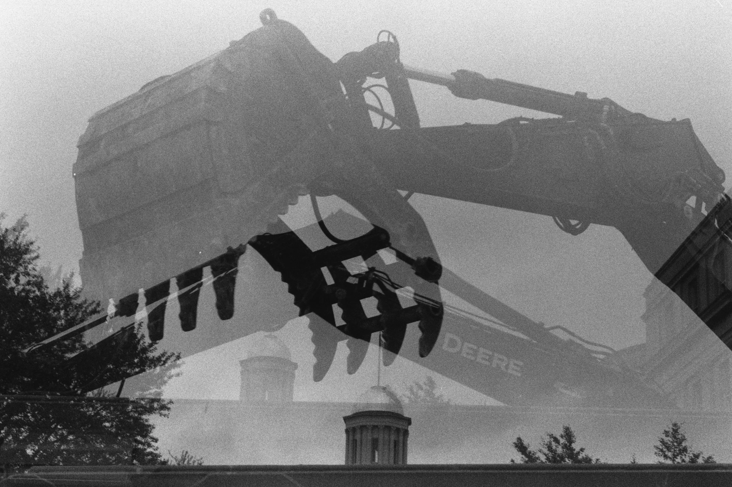 A black-and-white double exposure layers a Deere excavator’s bucket and arm over the Old Capitol dome and surrounding trees.