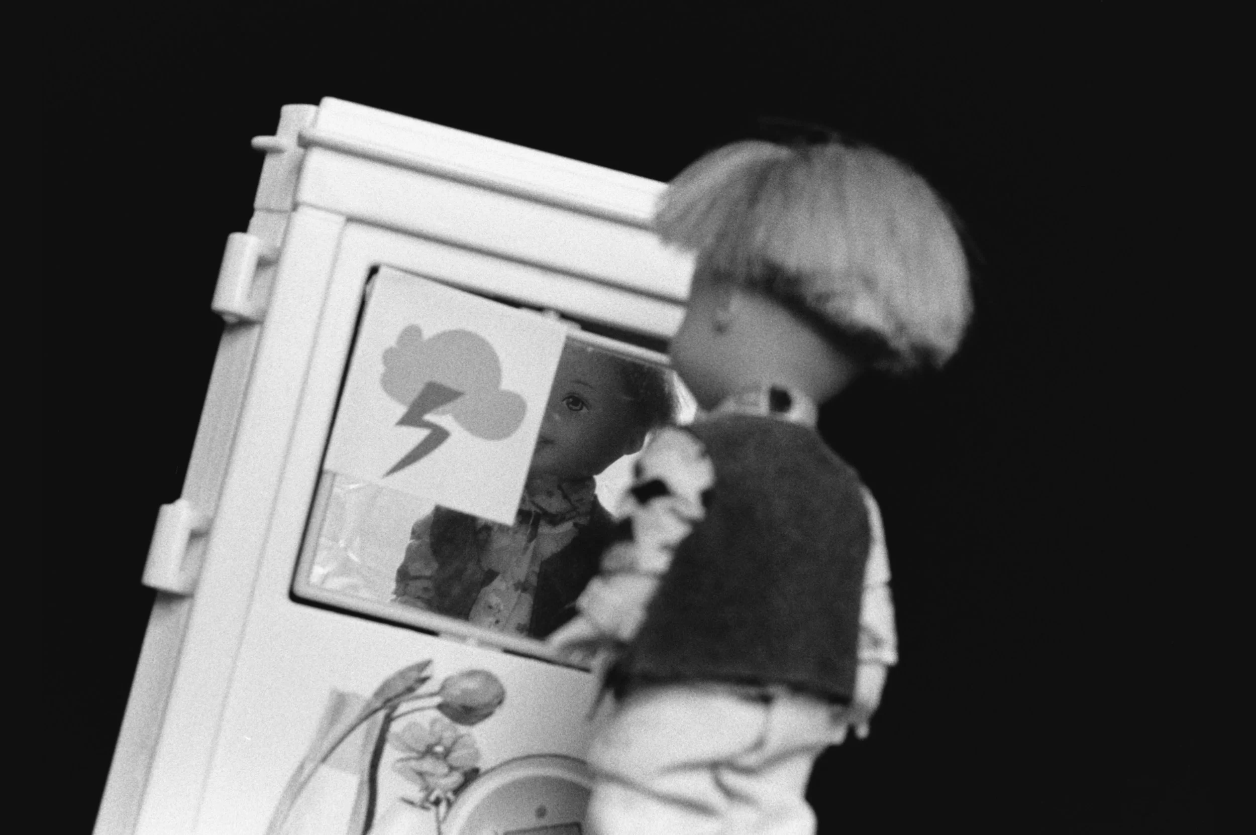A black-and-white photo shows Tommy, a bowl-cut doll, looking into a toy mirror, where his in-focus reflection is partly obscured by a storm cloud sticker.