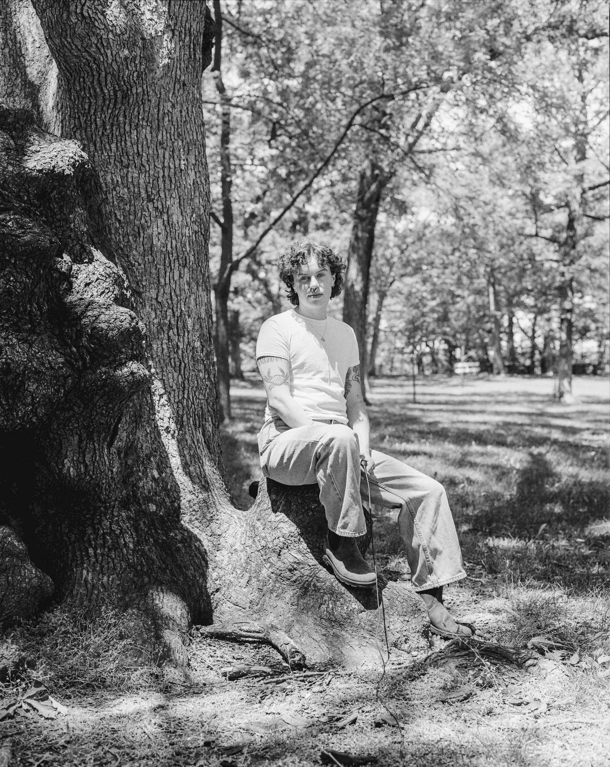 Liv sits on a tree trunk in a park in Iowa City. They look directly at the camera with a cable release in their hands. They have fair skin, curly short hair, and are wearing a white t-shirt, jeans, and boots.