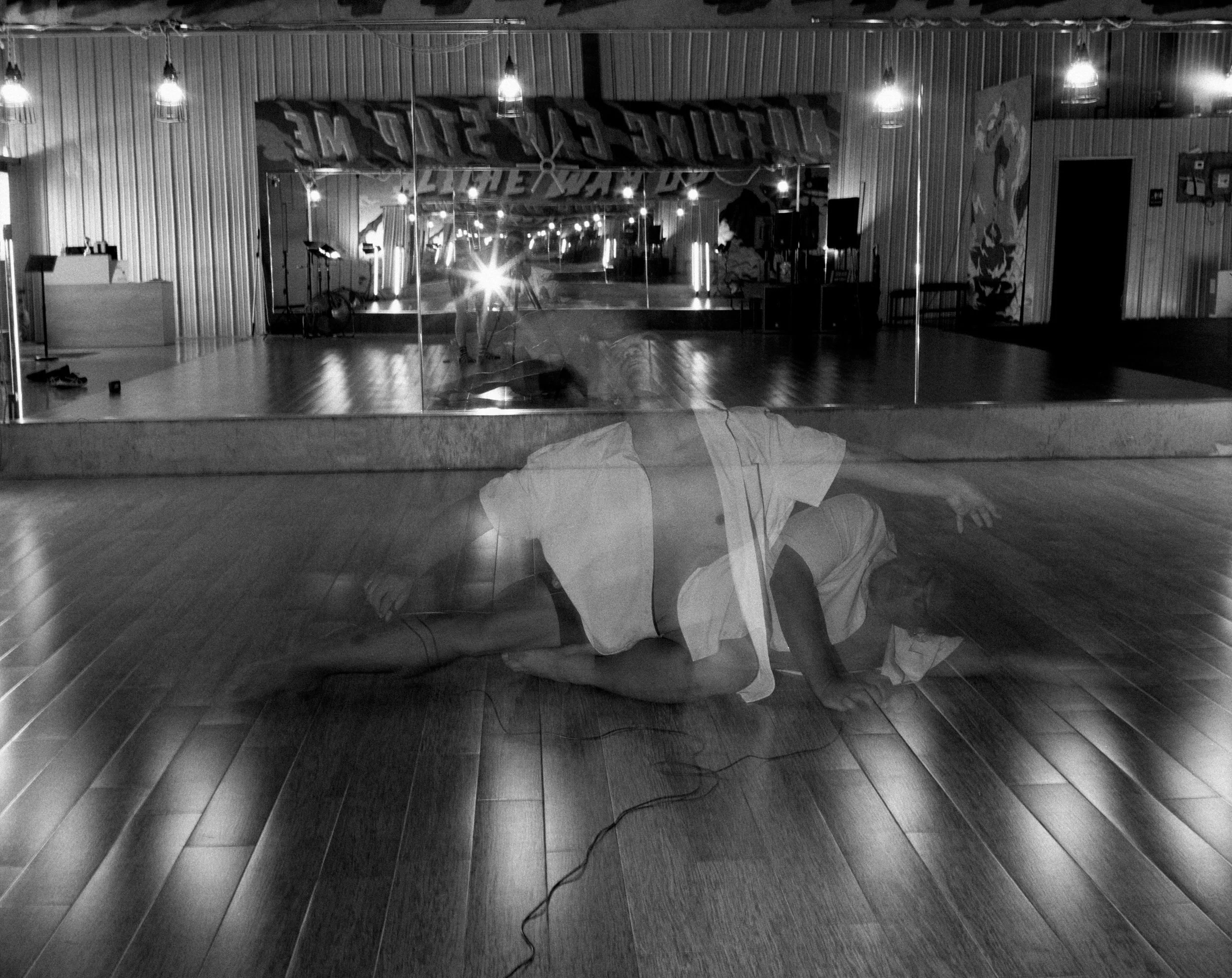 Miguel is on the floor of a dance studio. A double exposure through flash shows movement of his body. A mirror in the background reflects the flash.