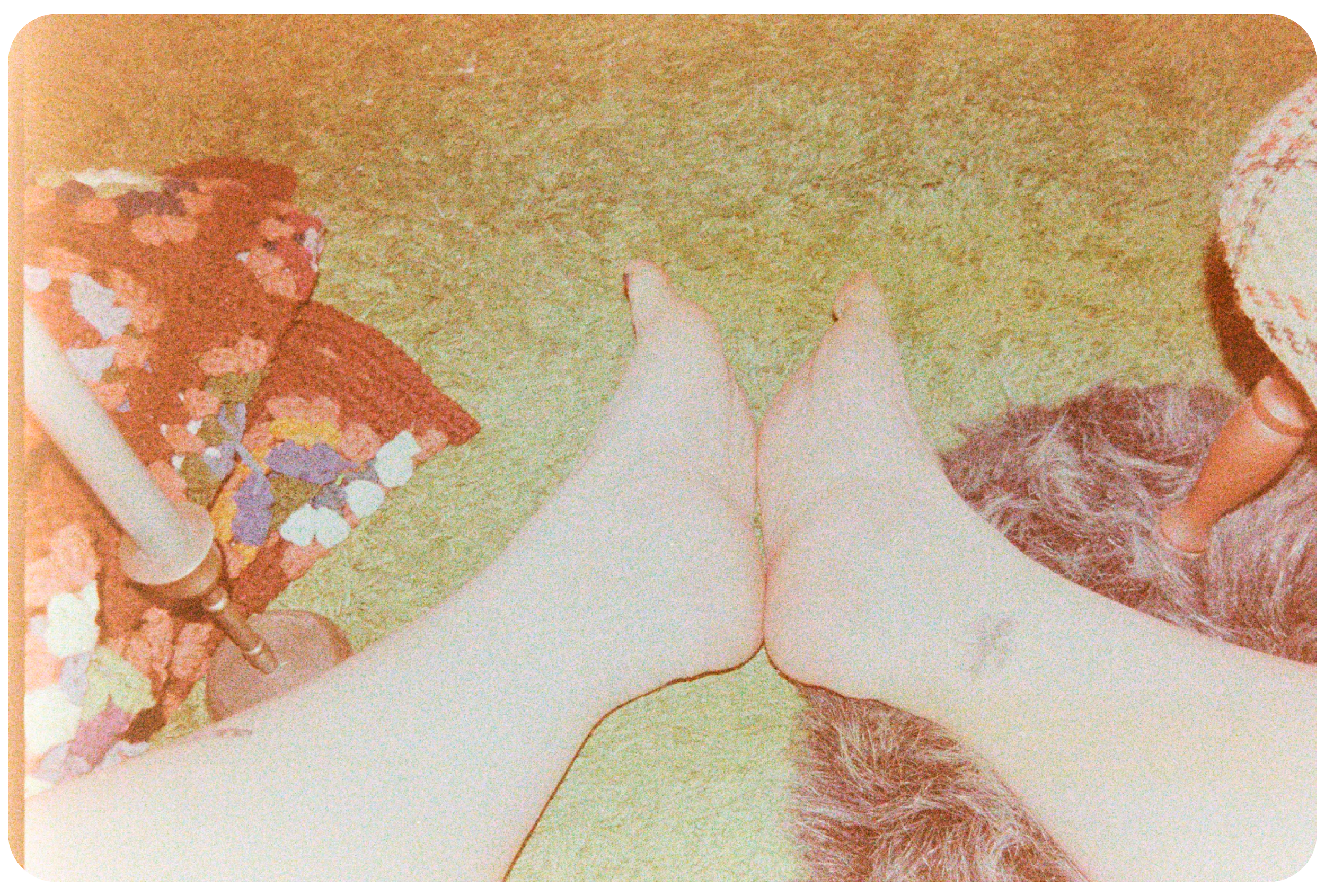 Top down view of a trans nonbinary person’s legs and feet, there is a clover tattoo on their right ankle. In the background there is a green rug, faux fur pelt, crocheted blanket, and a candle stick.