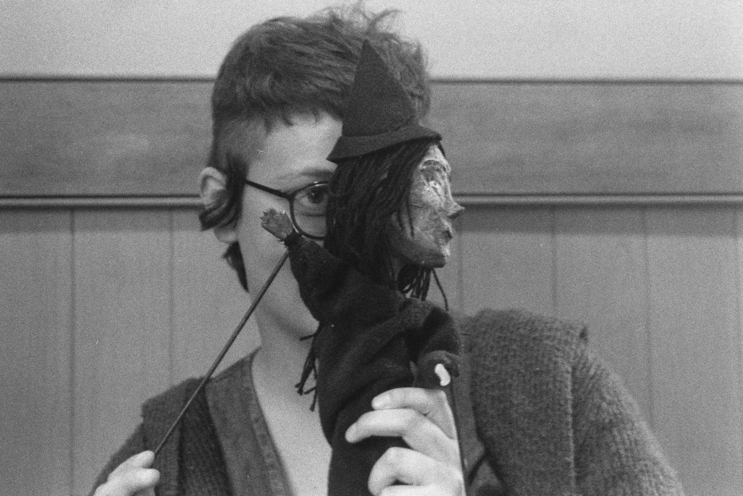 A black-and-white portrait shows a nonbinary person holding a witch doll over half their face as the doll raises a wooden hand.