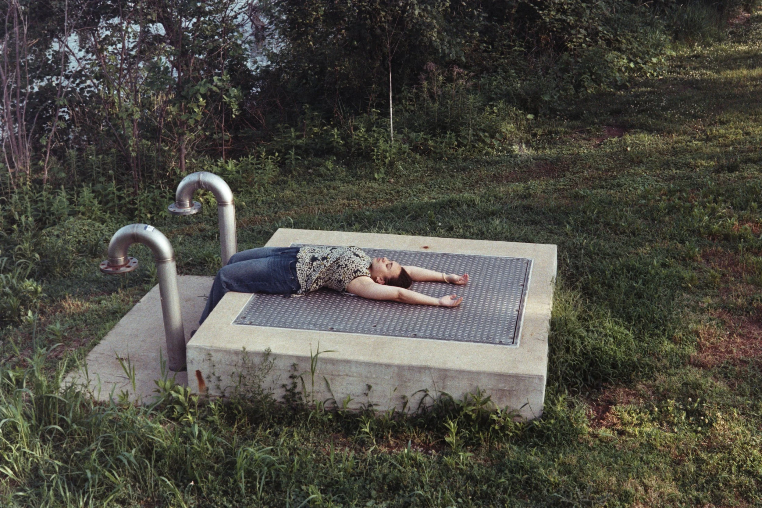 Pensée lies on a metal grate beside curved pipes by the Iowa City river, arms outstretched and eyes closed.