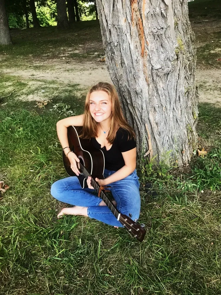 A young woman sitting cross-legged on grass beside a large tree, holding a black acoustic guitar, smiling at the camera during daytime in a wooded area.
