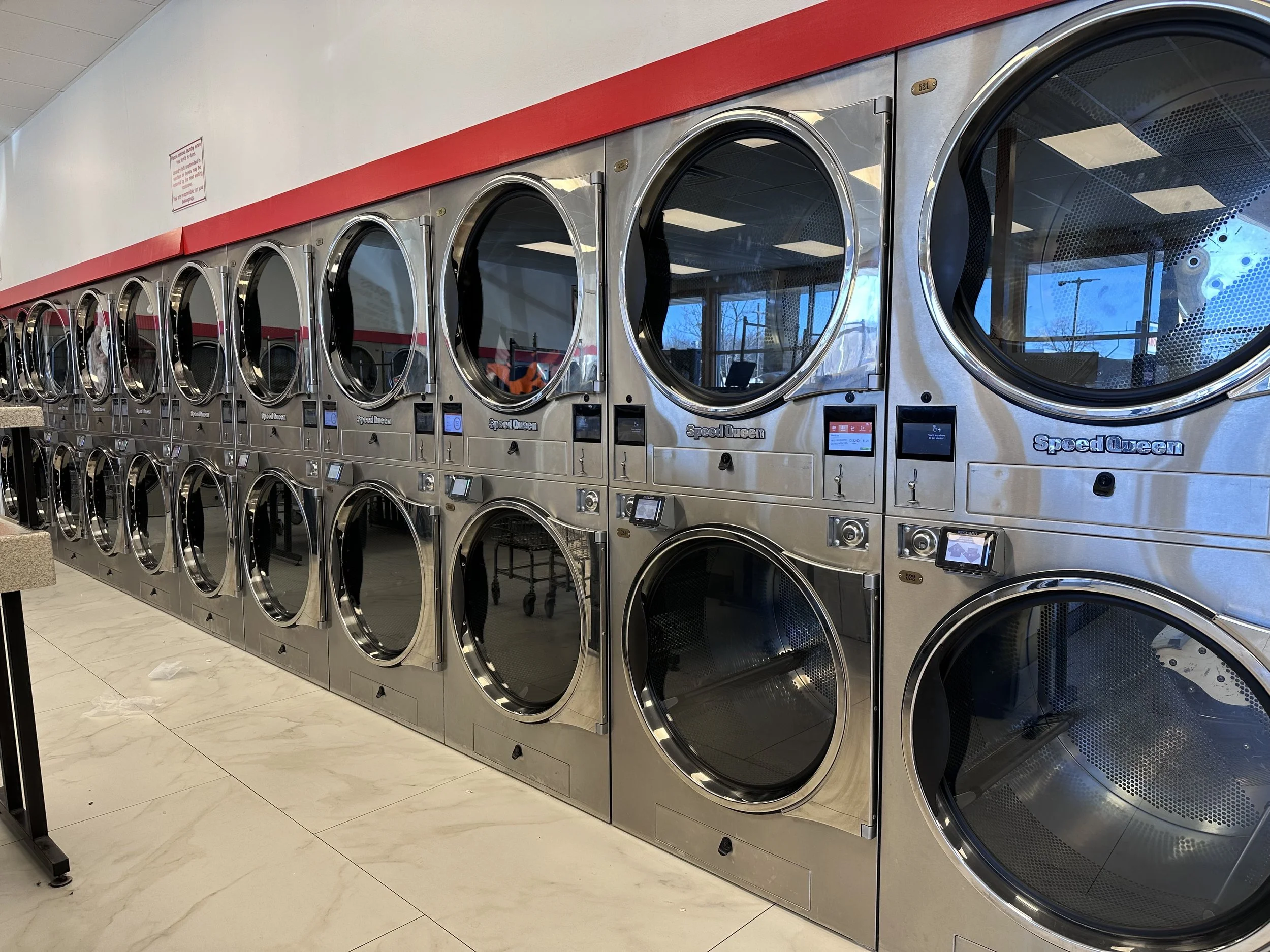 Row of commercial laundry dryers in a laundromat with digital controls and reflective stainless steel surfaces.