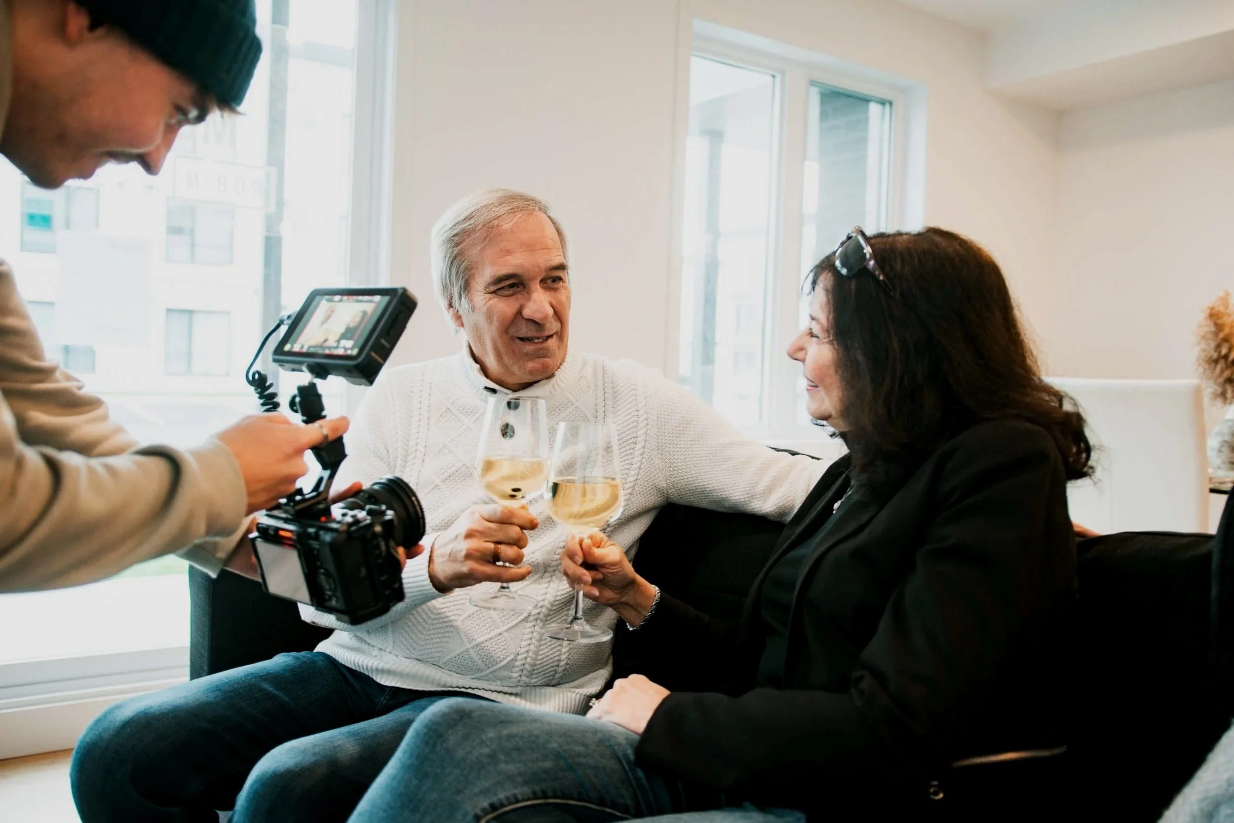 Deux personnes âgées souriantes levant des verres de vin pendant une séance de tournage vidéo à l'intérieur, avec un professionnel qui filme la scène.