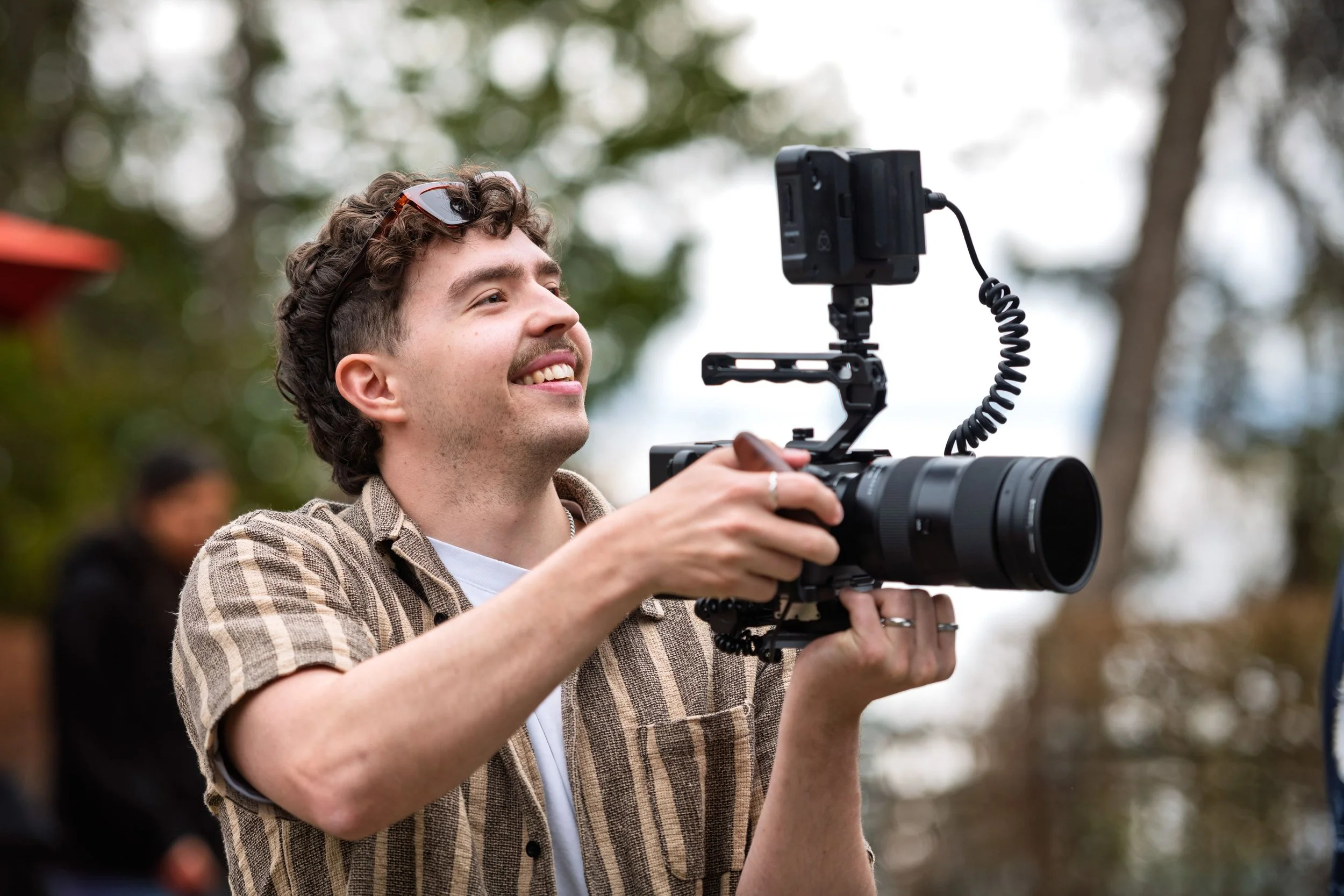 Vidéaste de H2 Productions, tenant une caméra avec stabilisateur, en extérieur avec des arbres en arrière-plan.