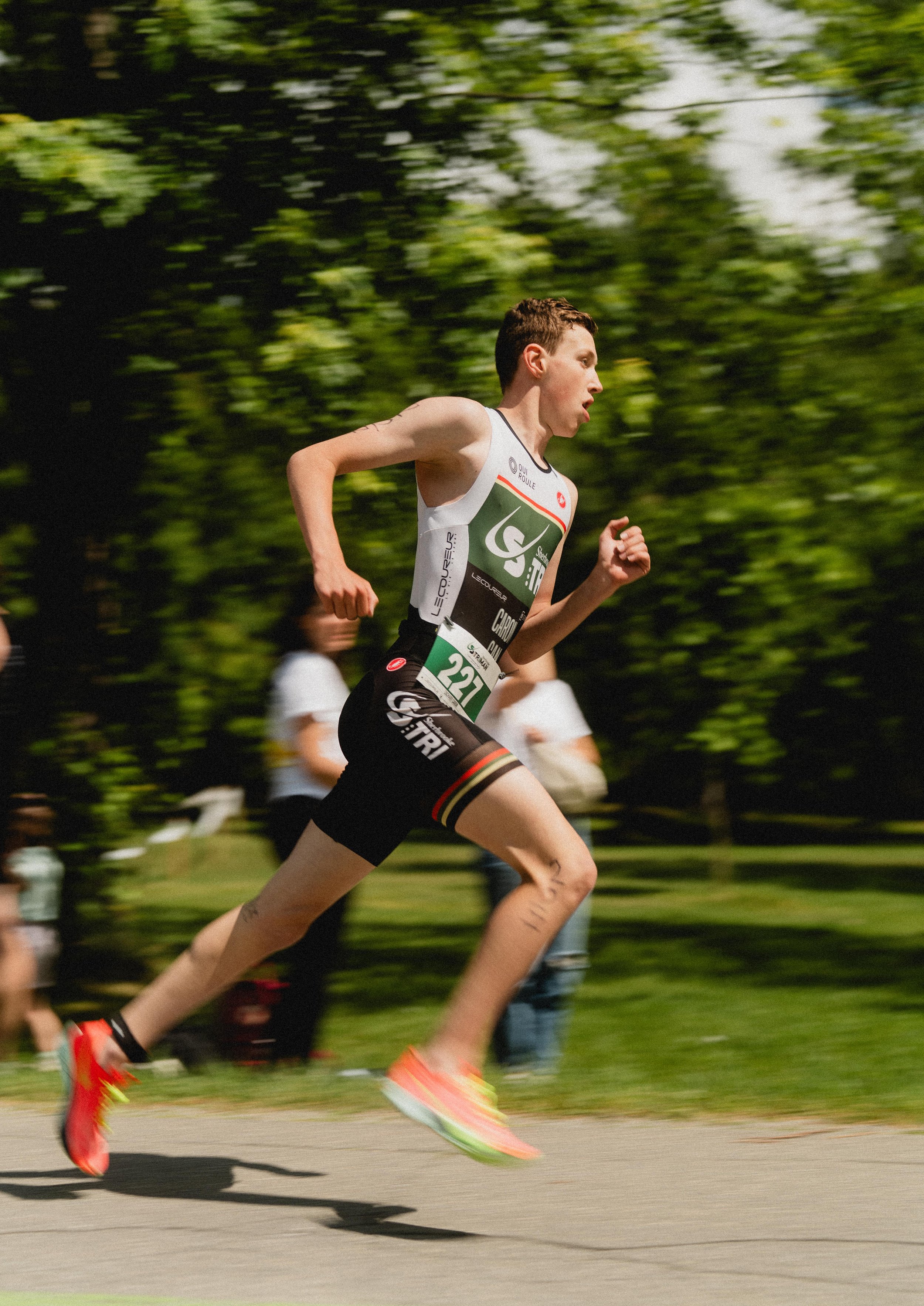 Un coureur participant à une course, en pleine accélération, portant un maillot et un short noirs avec des logos, chaussures de sport colorées, et un numéro 221, en extérieur avec des arbres en arrière-plan.