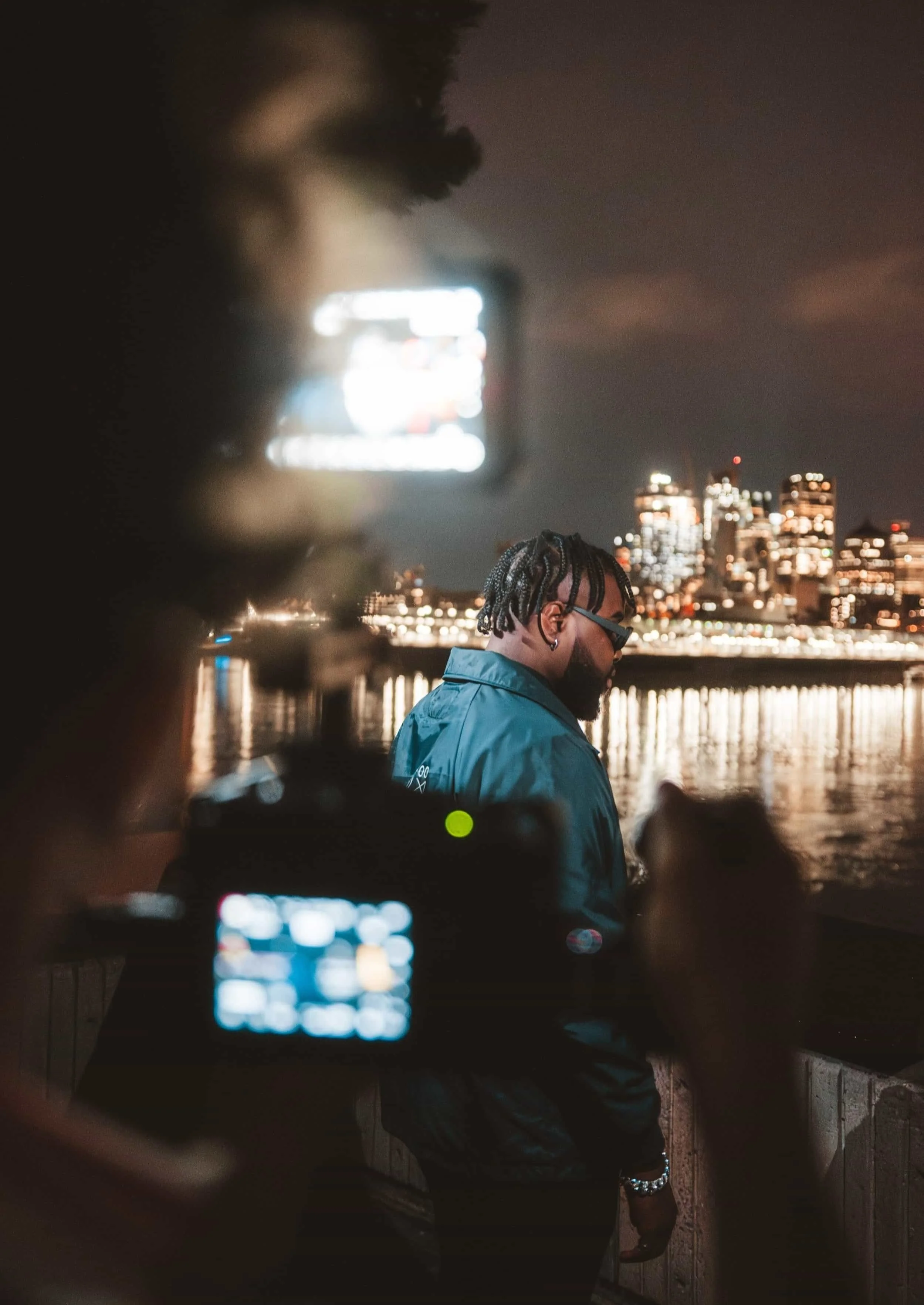 Un homme avec des dreadlocks et lunettes noires regarde de côté le centre-ville de Montréal, illuminée la nuit, photographié pendant un vidéoclip à travers un appareil photo avec écran lumineux