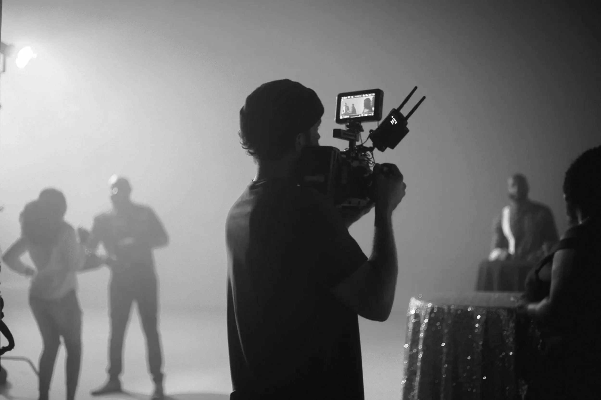 A person operating a video camera films a scene with multiple models standing in a line, some with hands on hips, in a studio setting. The image is black and white.