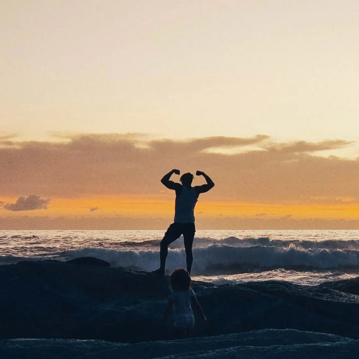 A father strikes a funny pose in front of his kid in Cape Town at sunset.