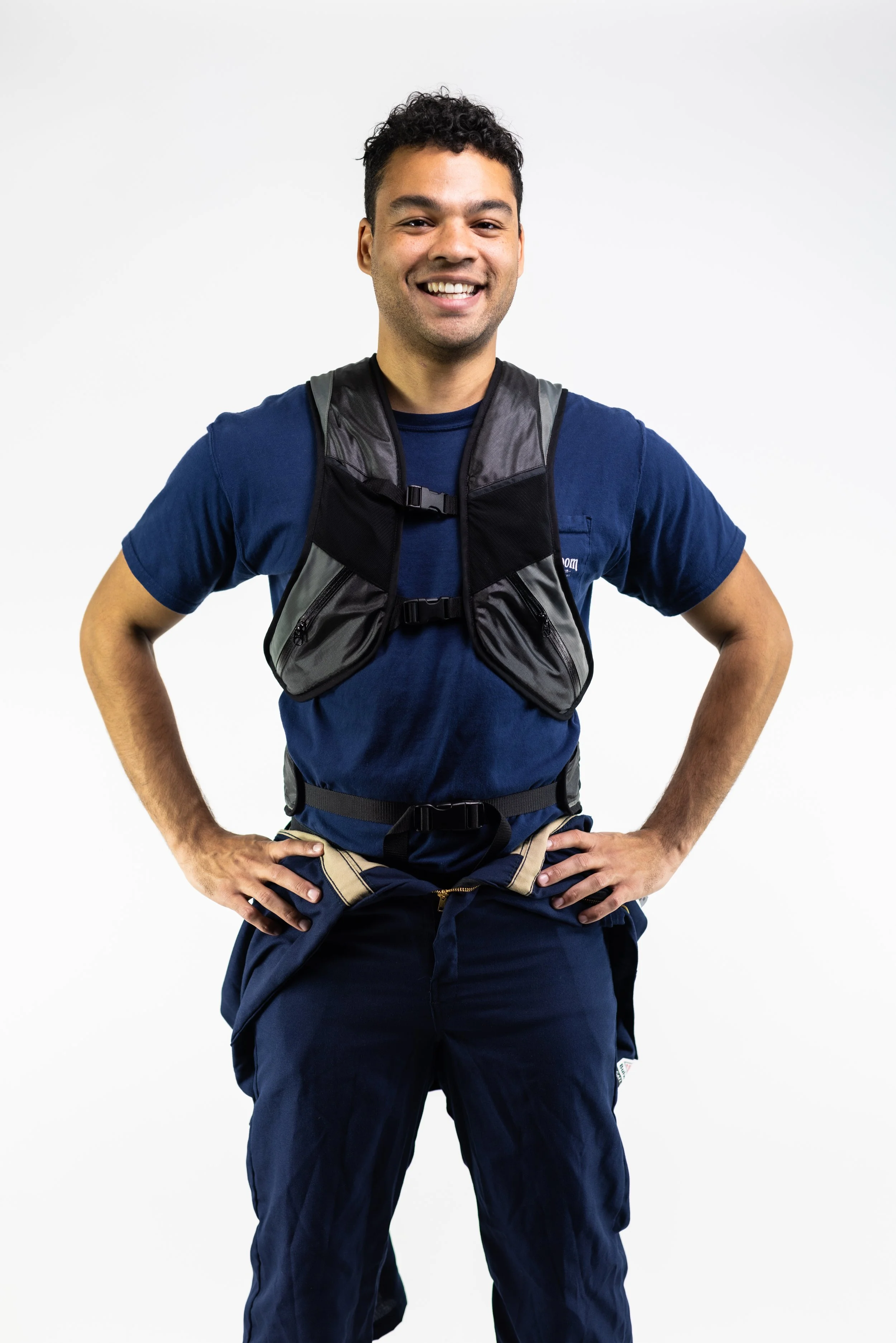 Young man standing with hands on hips, smiling, wearing a navy blue t-shirt, navy blue pants, and a black hydration vest, against a plain white background.
