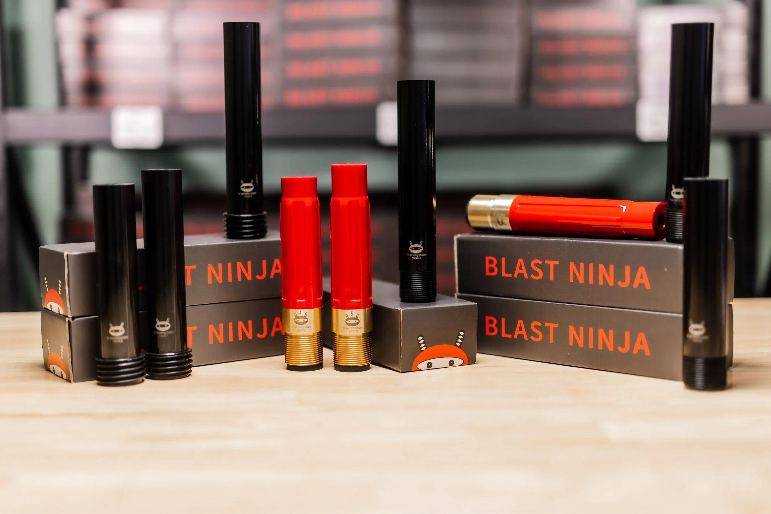 Display of Blast Ninja fireworks with black and red tubes on black boxes featuring the Blast Ninja logo are arranged on a wooden surface, with blurred shelves in the background.