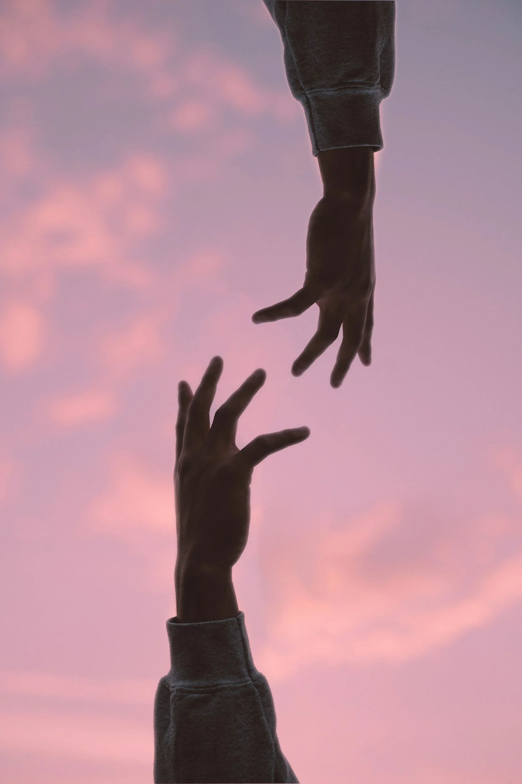 Two hands reaching upward into a pastel pink and purple sky, with one hand holding the other lightly from above.