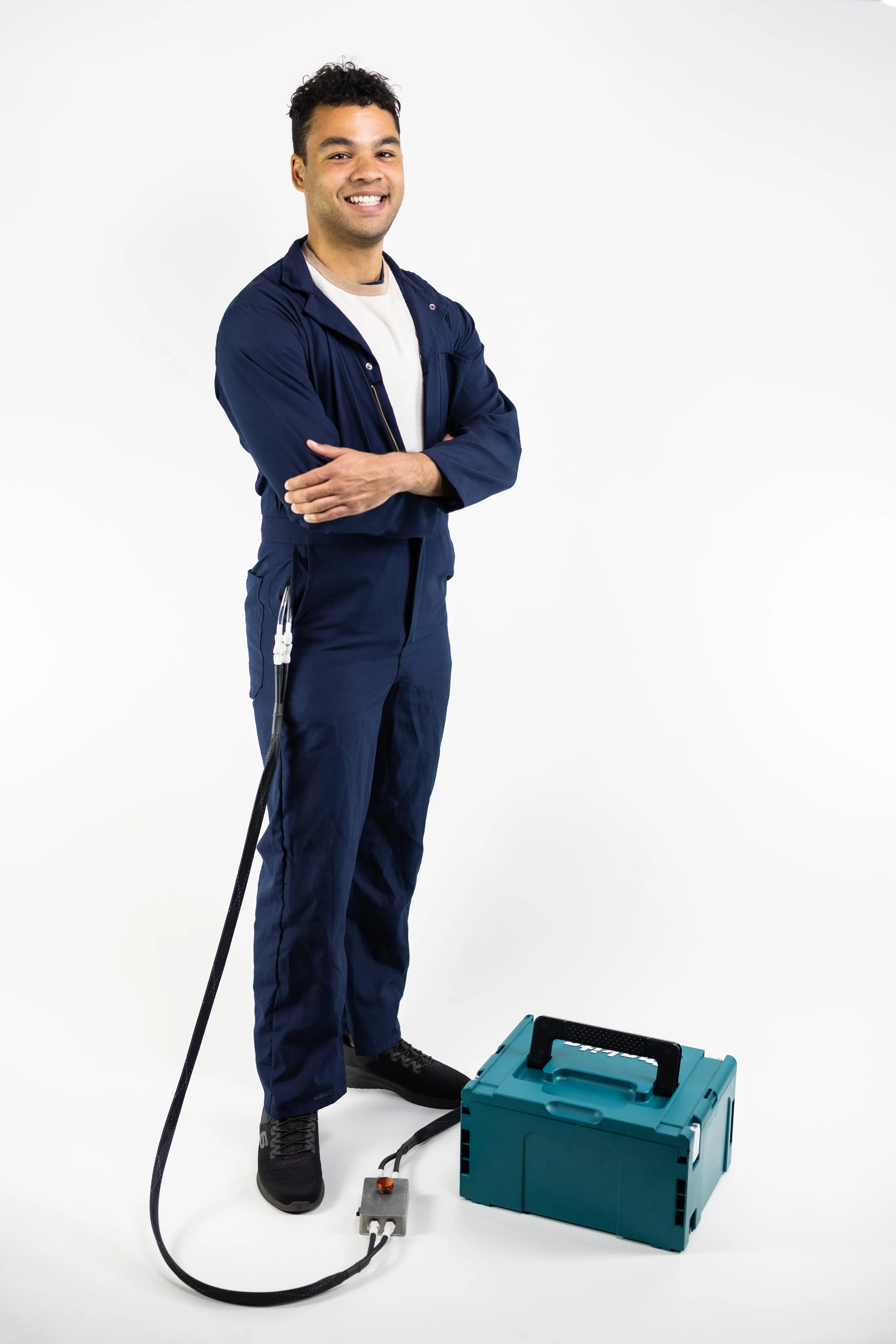 A man dressed in dark blue work clothes standing on a white background with a portable blue generator on his left, a power cord running from the generator, and some connected equipment on a gray platform.