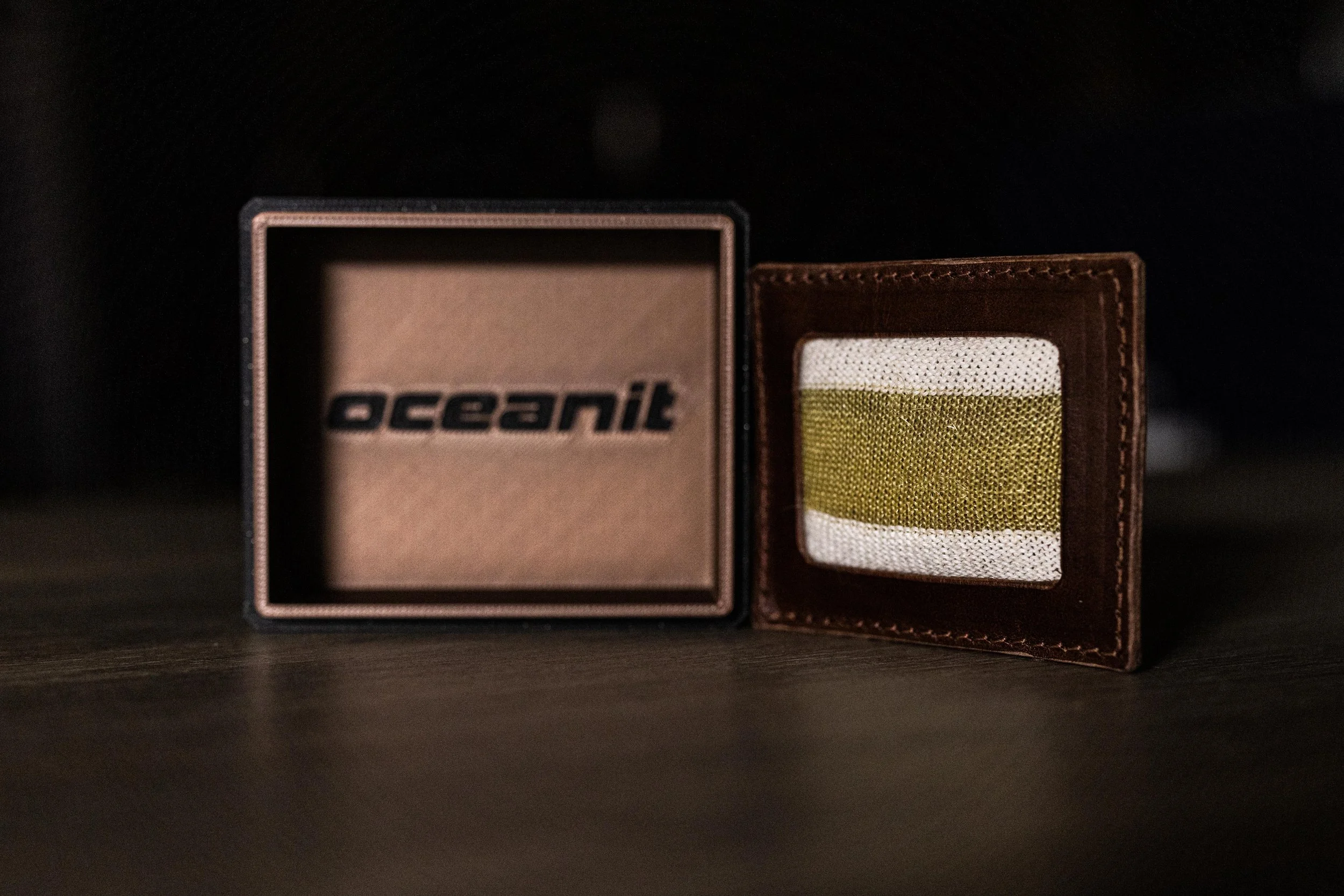 A brown leather wallet and a small box with the word 'Oceanit' on it, placed on a dark surface with a dark background.