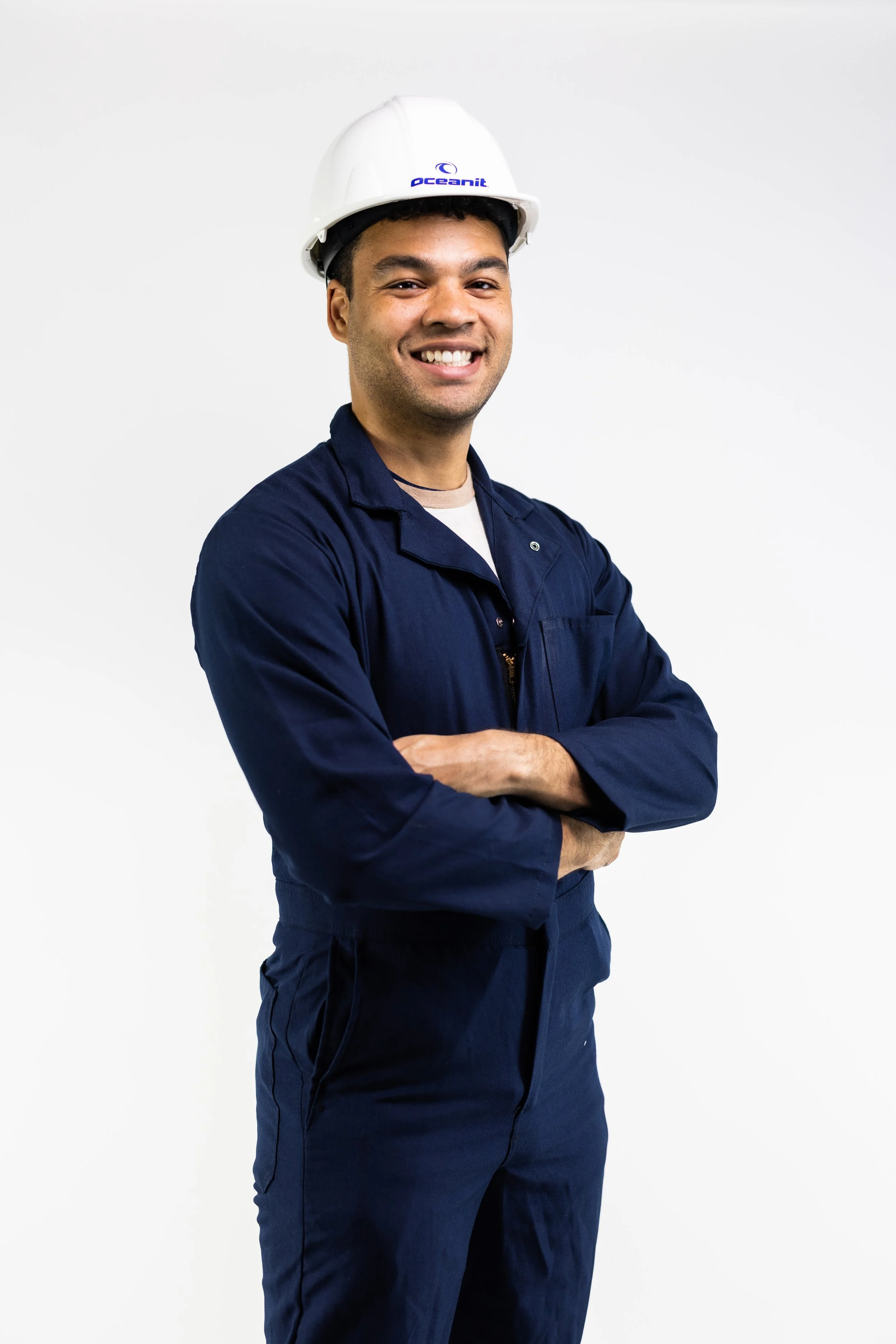 A smiling man in a navy blue work uniform and white safety helmet with a logo, standing with arms crossed against a plain white background.