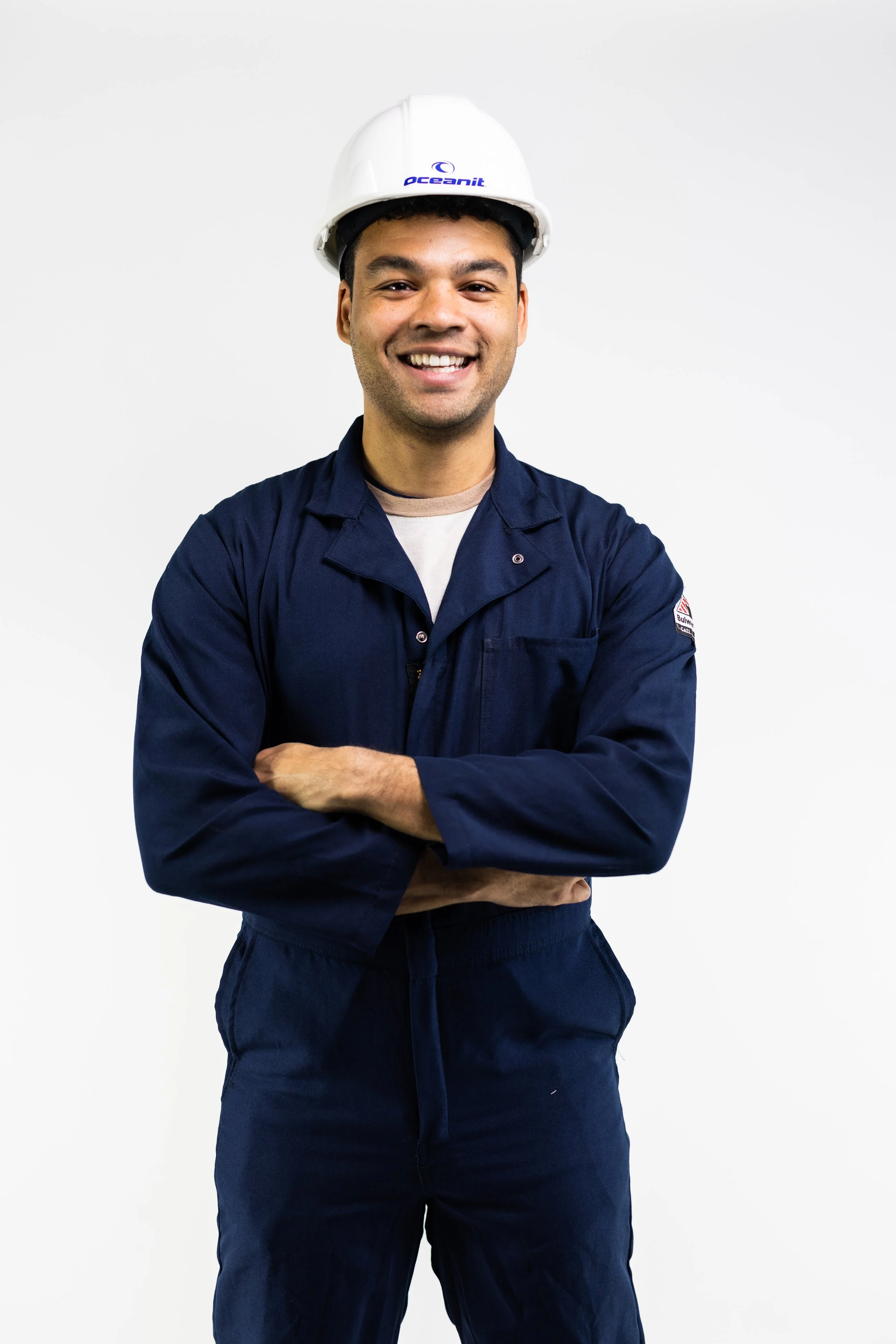 A smiling man wearing a white safety helmet with the logo 'Oceanit' and a navy blue work uniform with a patch on the sleeve, standing against a plain white background.
